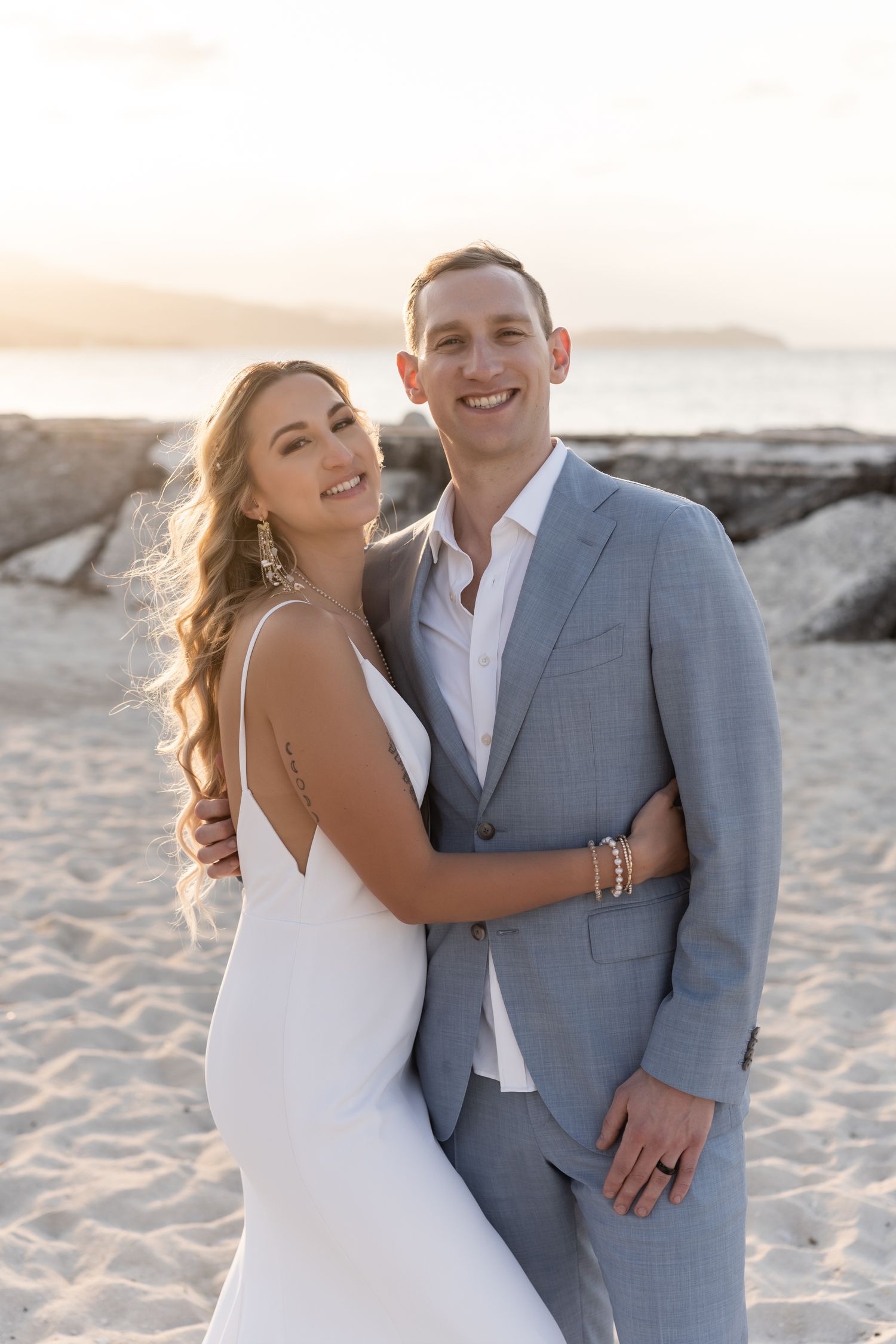 Newlyweds embracing on beach at sunset in elegant wedding attire.