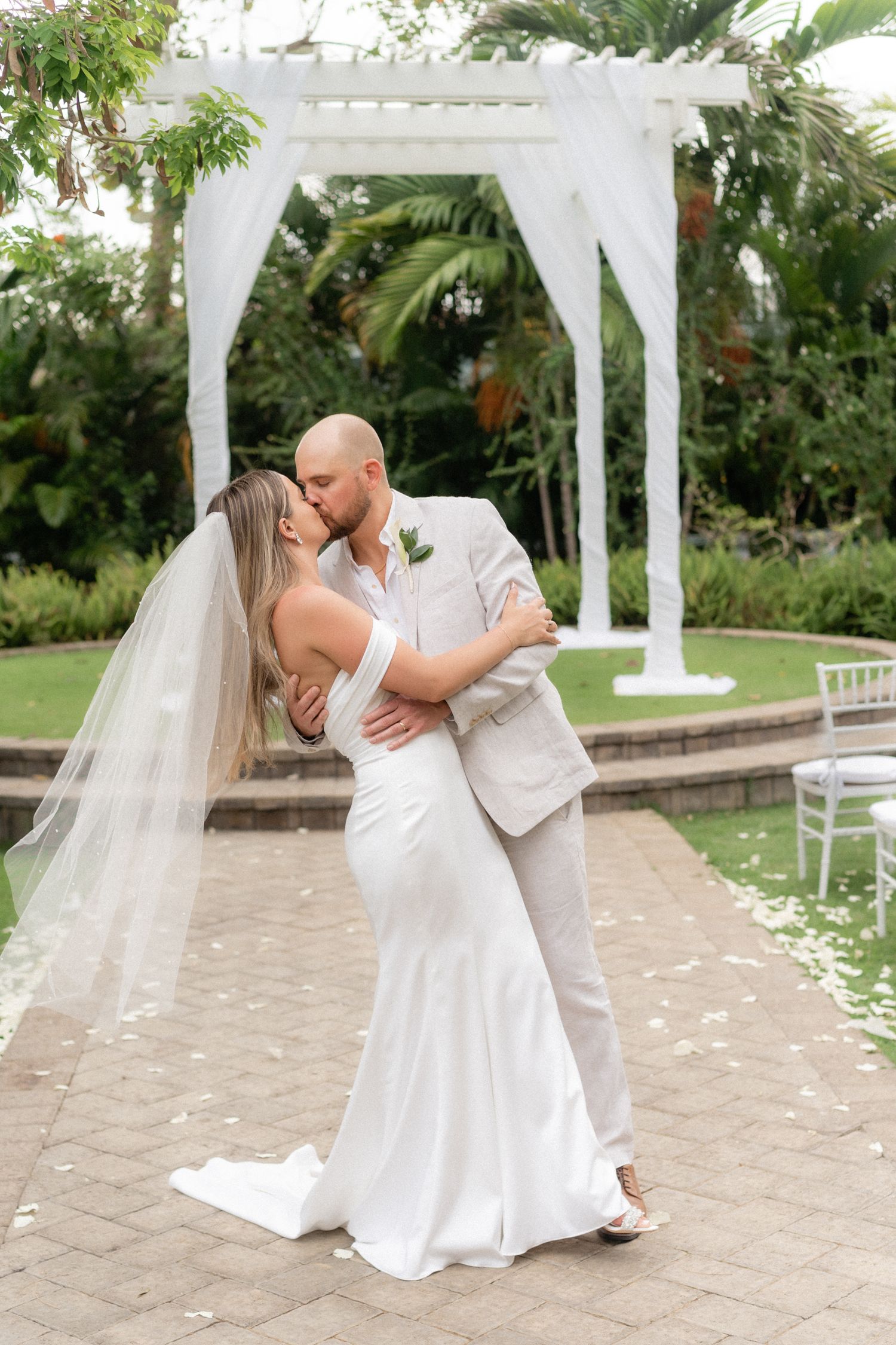 Newlyweds embrace under white draped pergola on stone paved garden pathway.