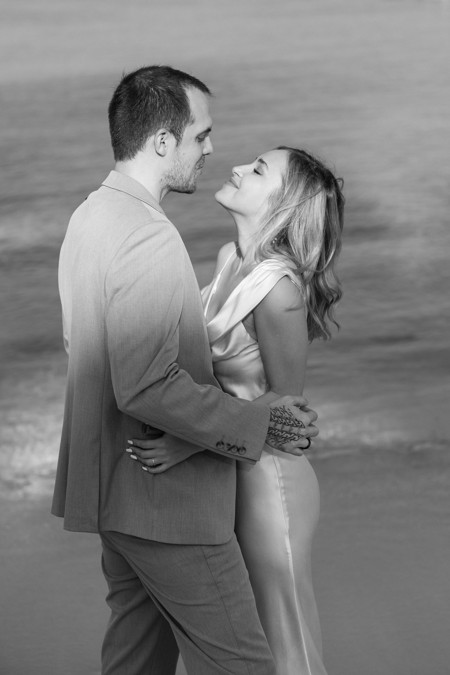 A romantic black and white portrait of a couple in formal attire embracing on the beach.