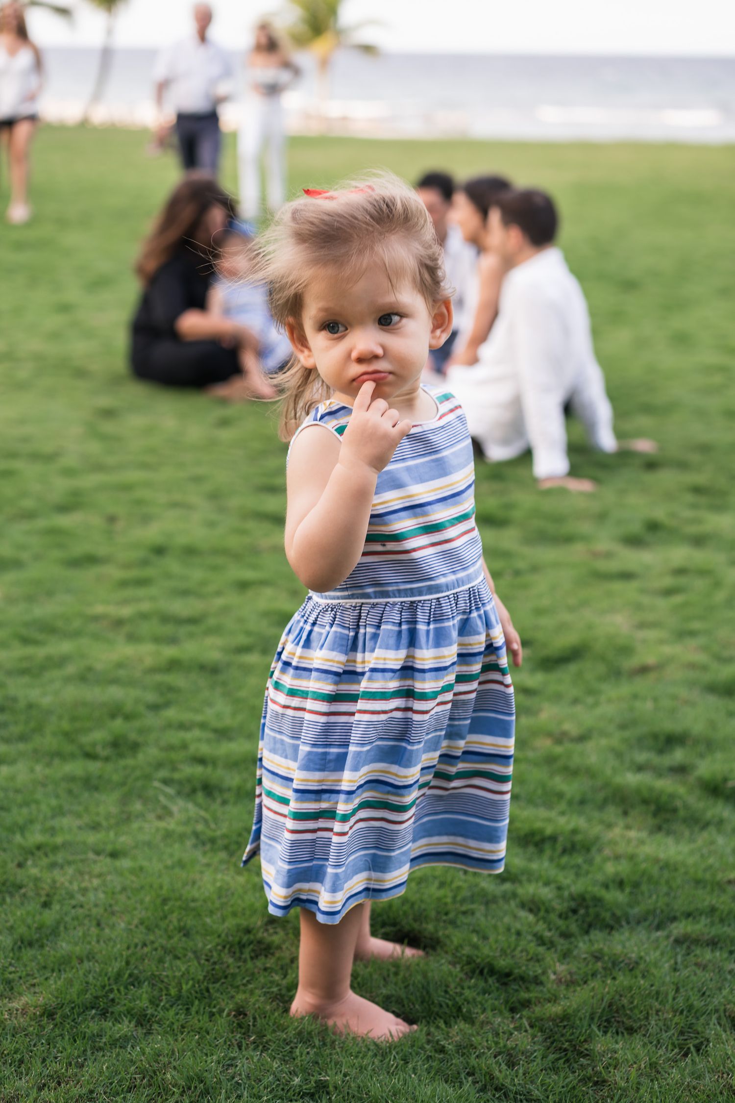 Child in striped dress standing barefoot on grass with others seated in background.