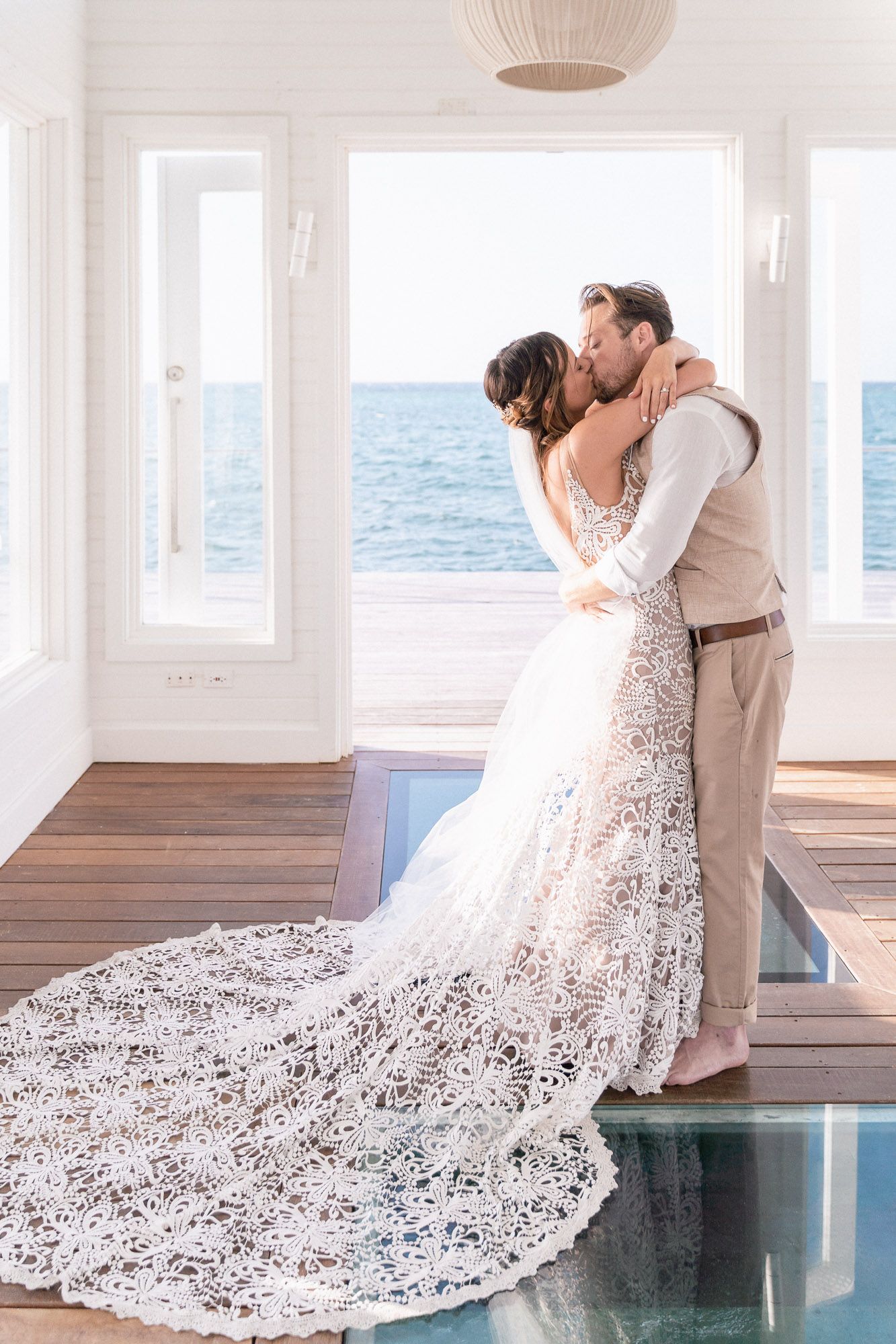A romantic kiss between newlyweds in a white seaside venue with floor-length lace wedding dress.