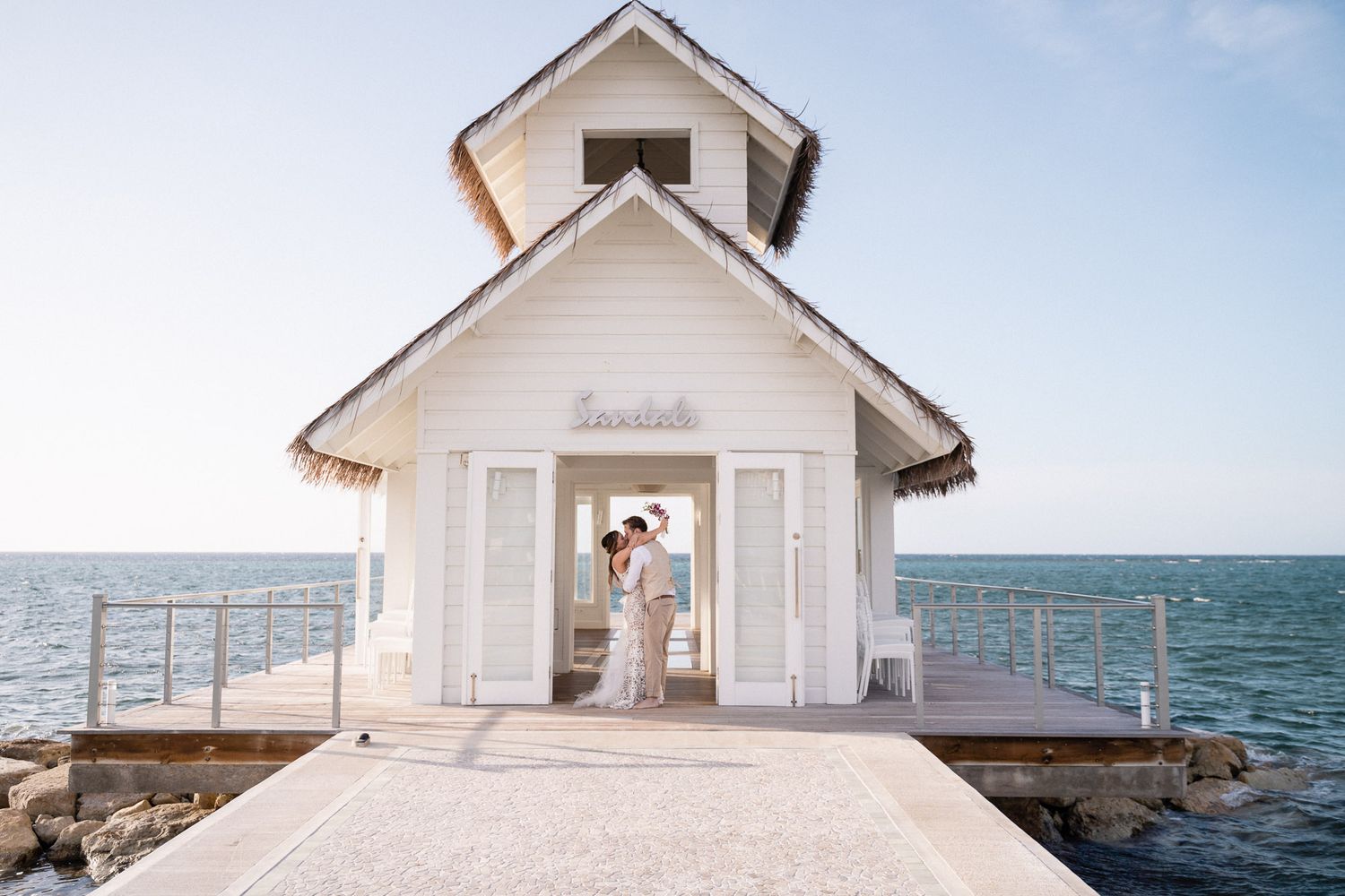 A white seaside wedding chapel with decorative trim sits on a pier overlooking the ocean on a sunny day.