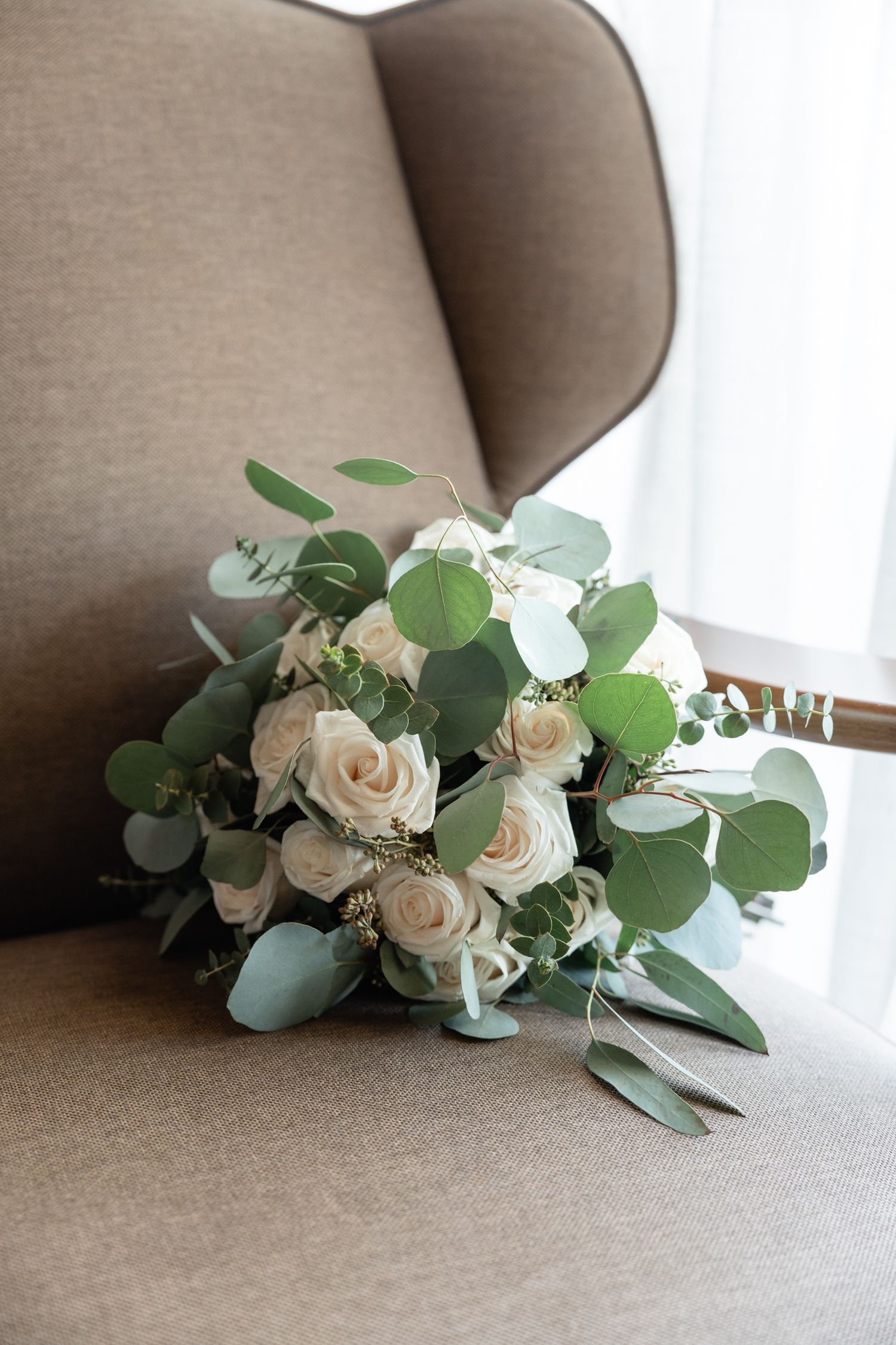 Elegant white rose and eucalyptus bridal bouquet resting on beige armchair.