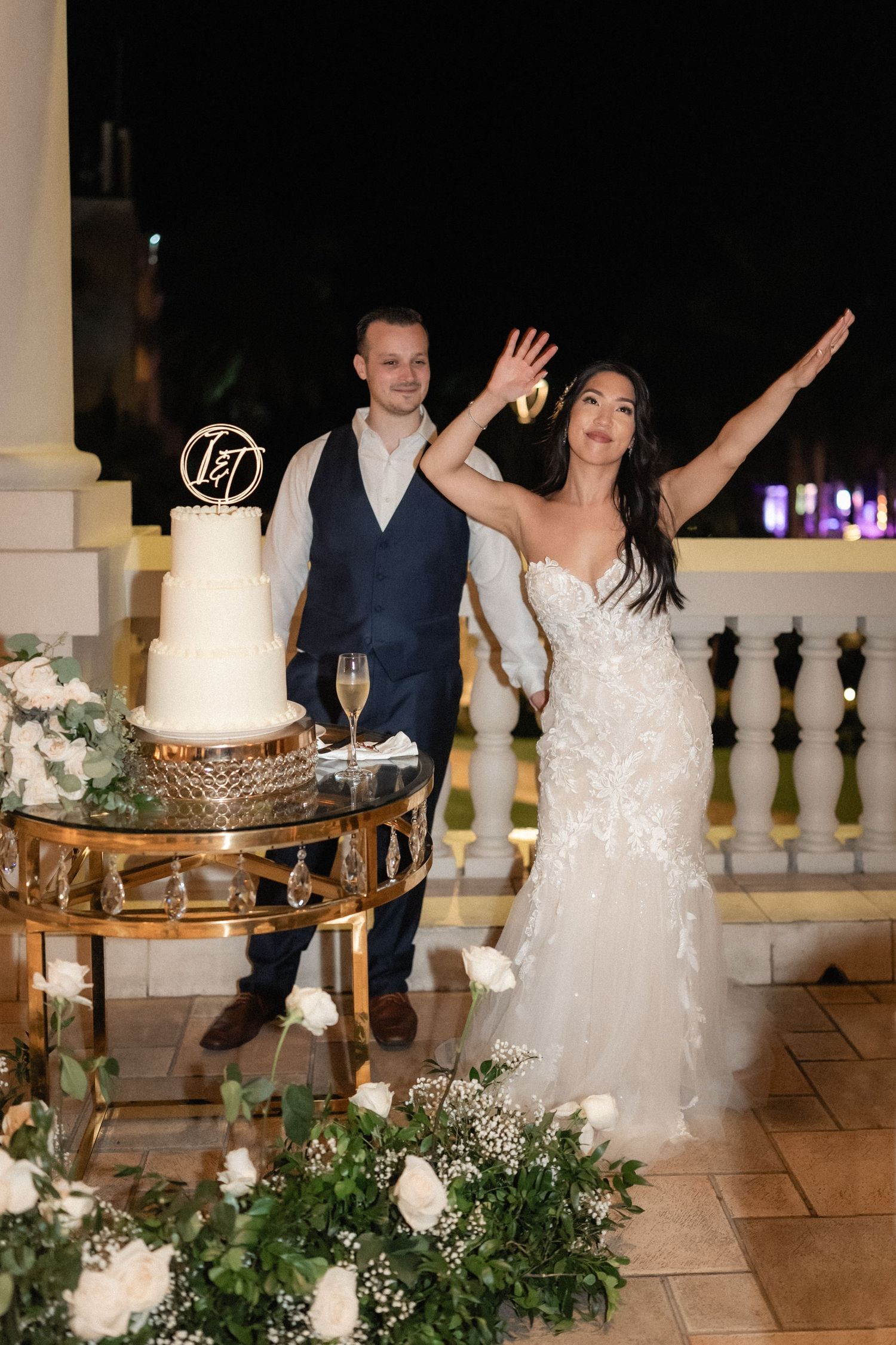 Couple poses with enthusiasm next to their elegant white wedding cake adorned with floral decorations.