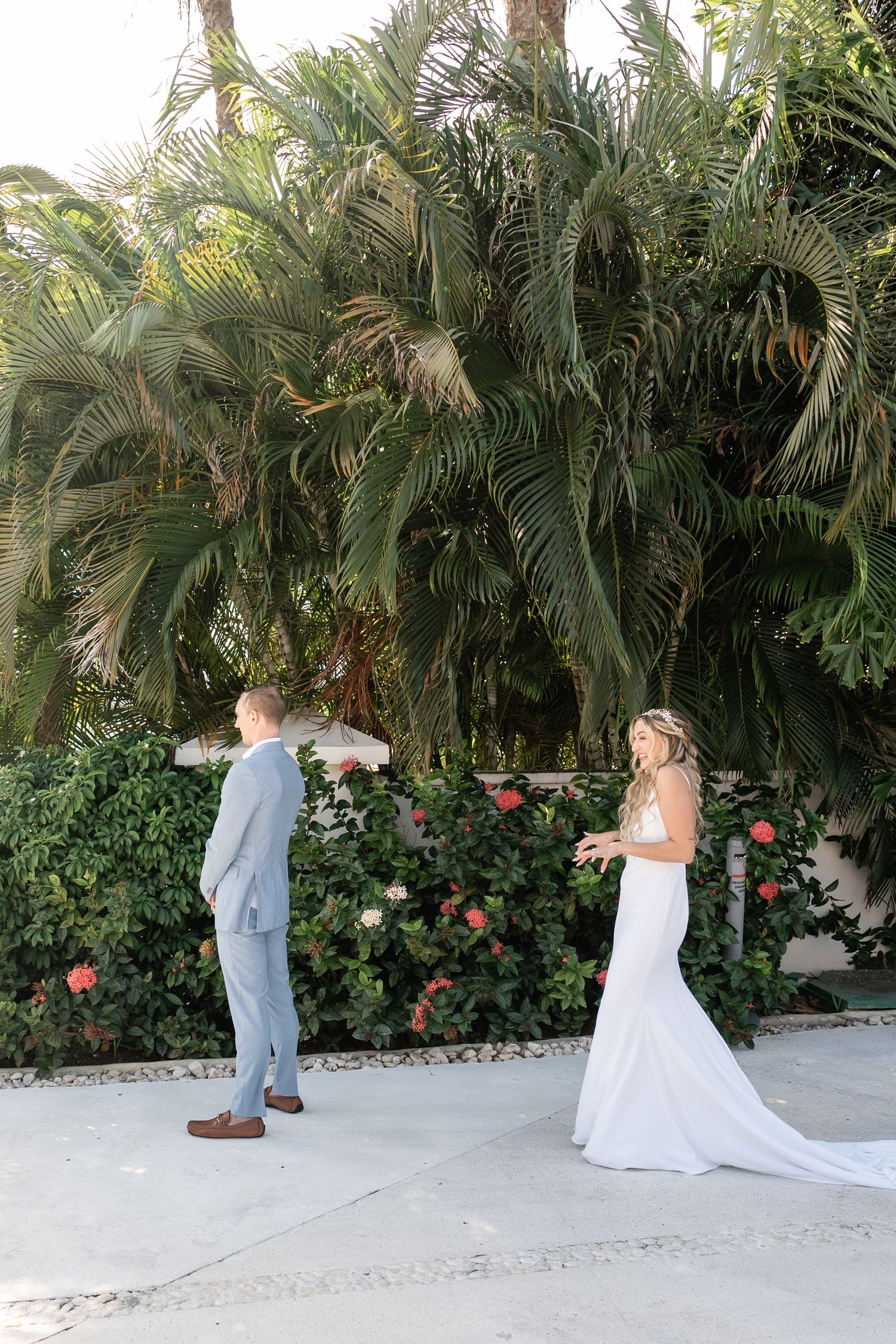 A couple in wedding attire sharing their first look moment against a backdrop of tropical palm trees and roses.