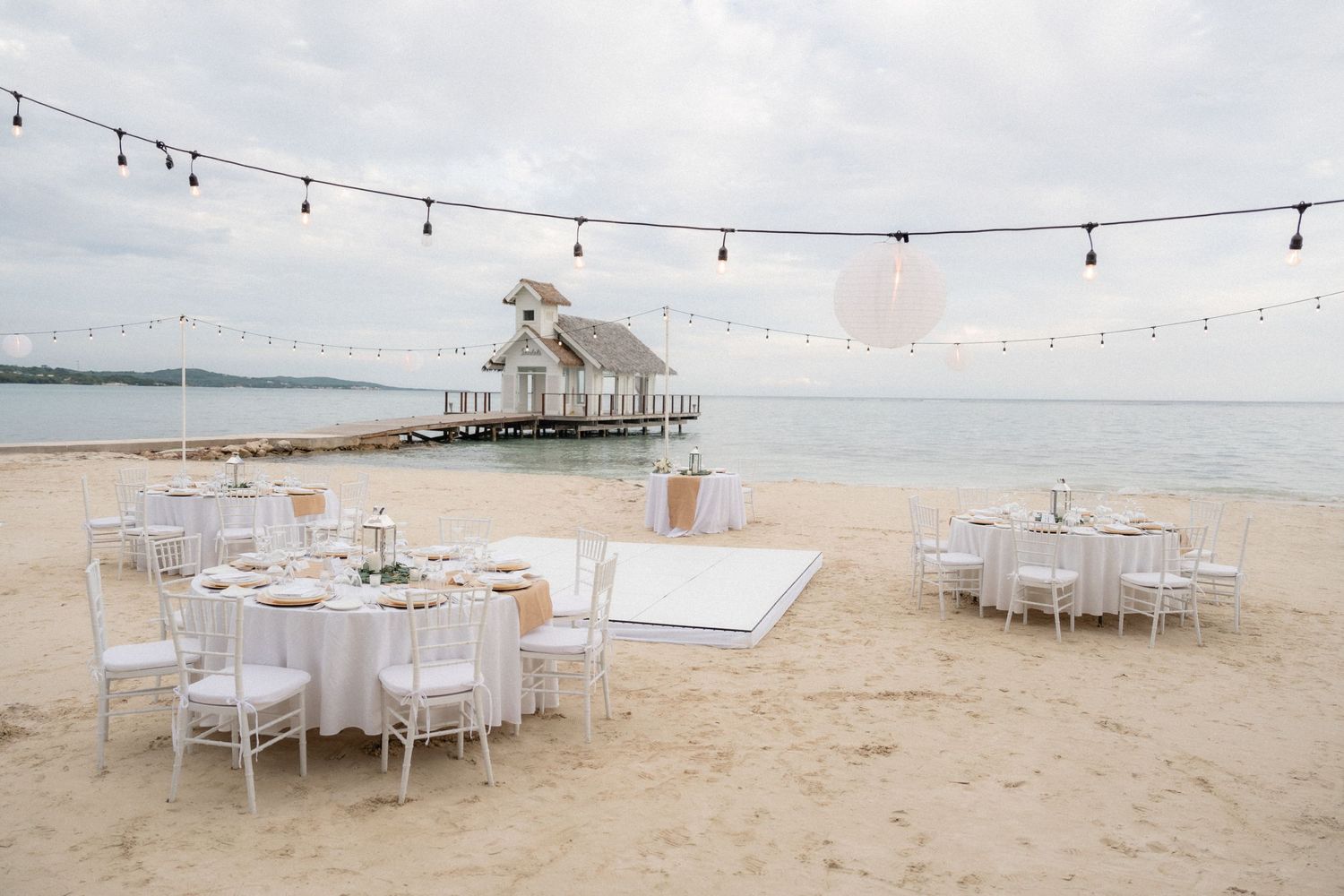 Romantic beach wedding setup with string lights and white tables near an ocean pier pavilion.
