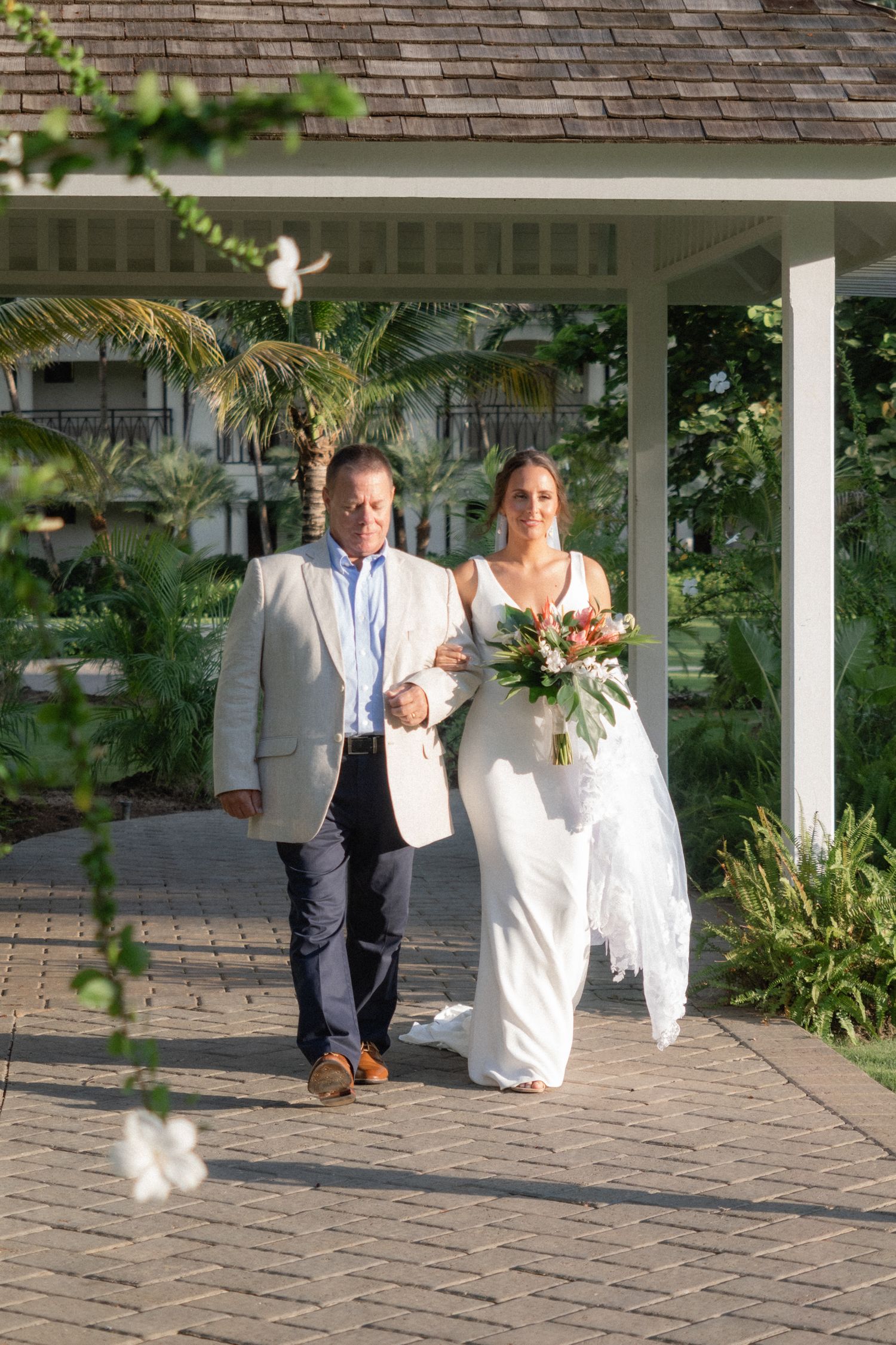 Wedding processional on a brick pathway under a covered walkway with tropical landscaping.