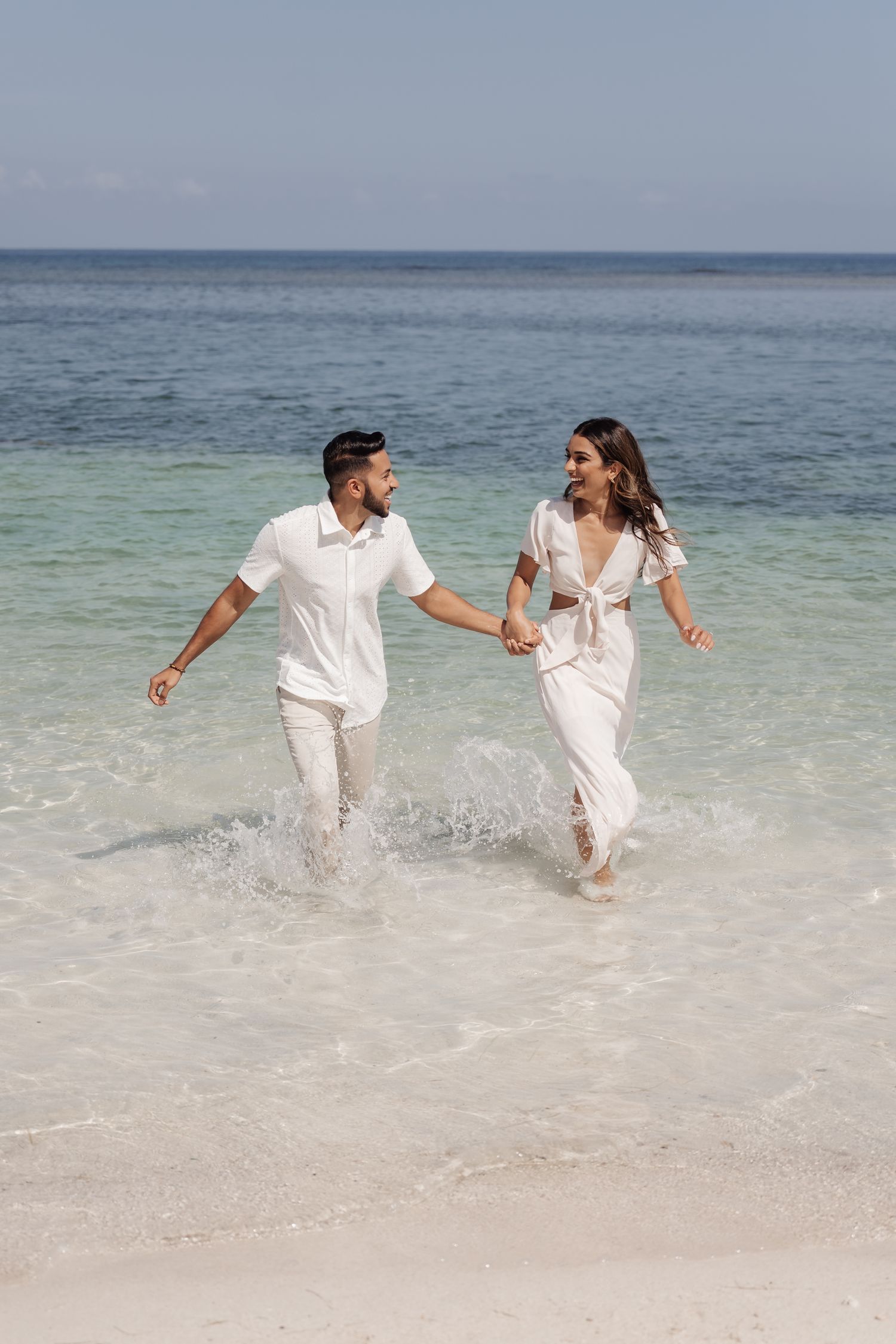 A couple in white outfits running joyfully through shallow ocean waters on a tropical beach.