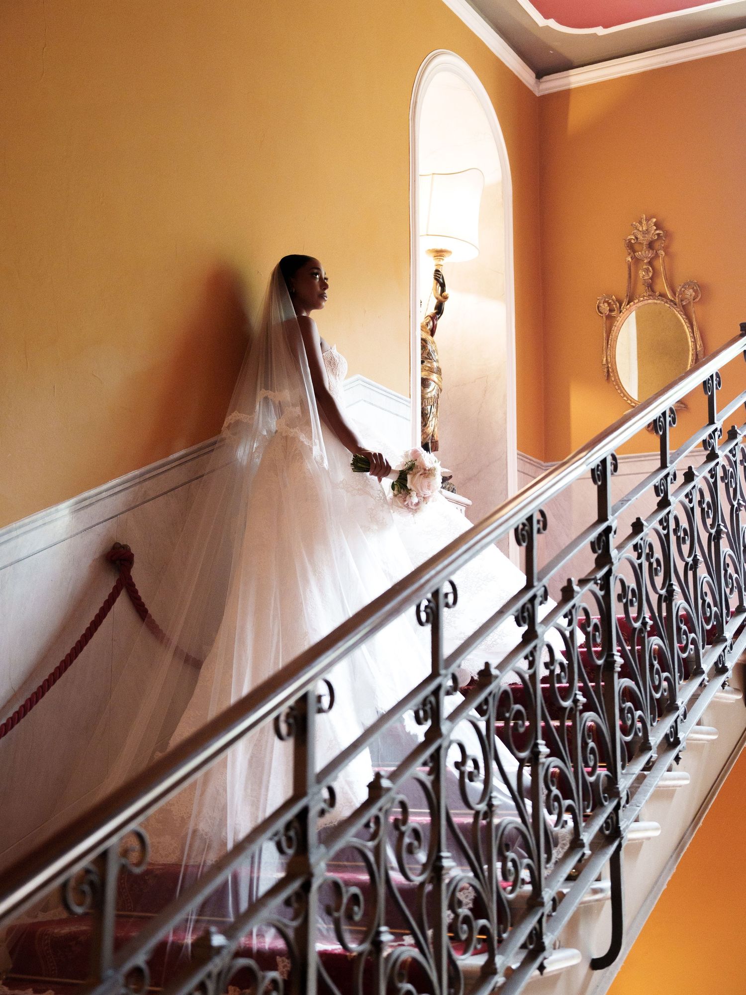 A flowing white dress trails up an ornate staircase with wrought iron railings and orange walls.