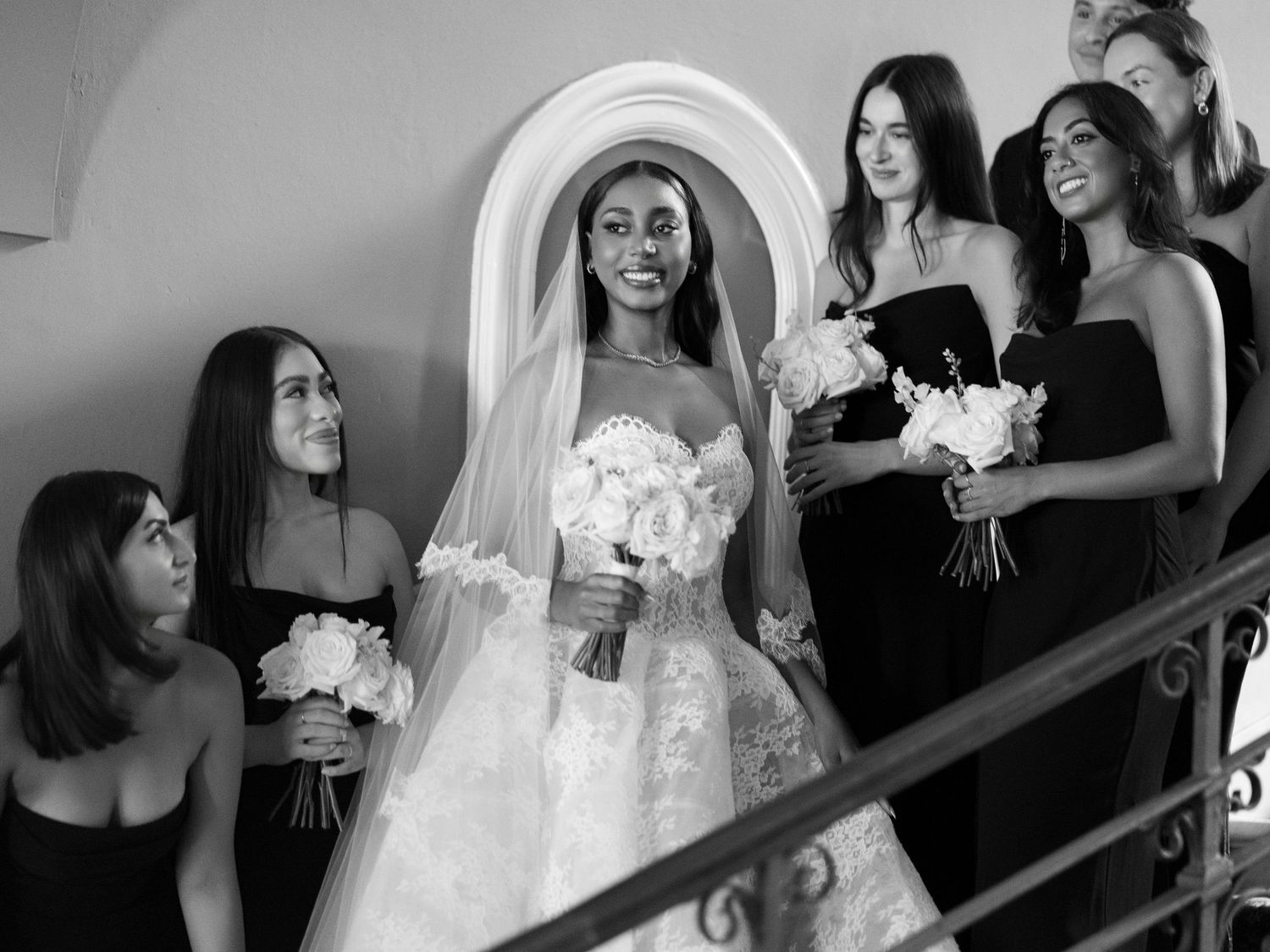 Black and white wedding photo showing a bride in white gown with bridesmaids in dark dresses on a staircase.