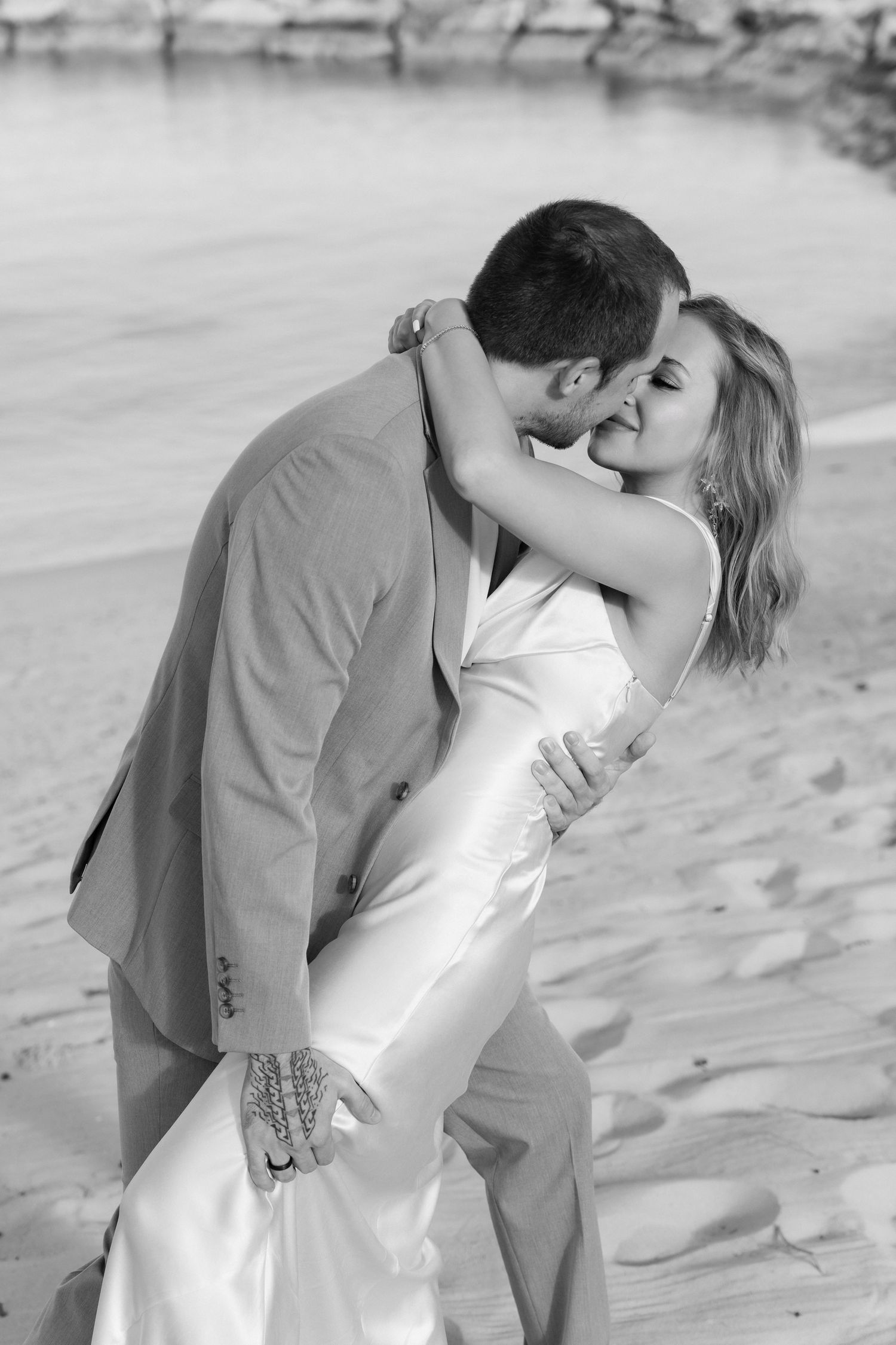 Black and white photograph of newlyweds embracing in an intimate kiss on a sandy beach.