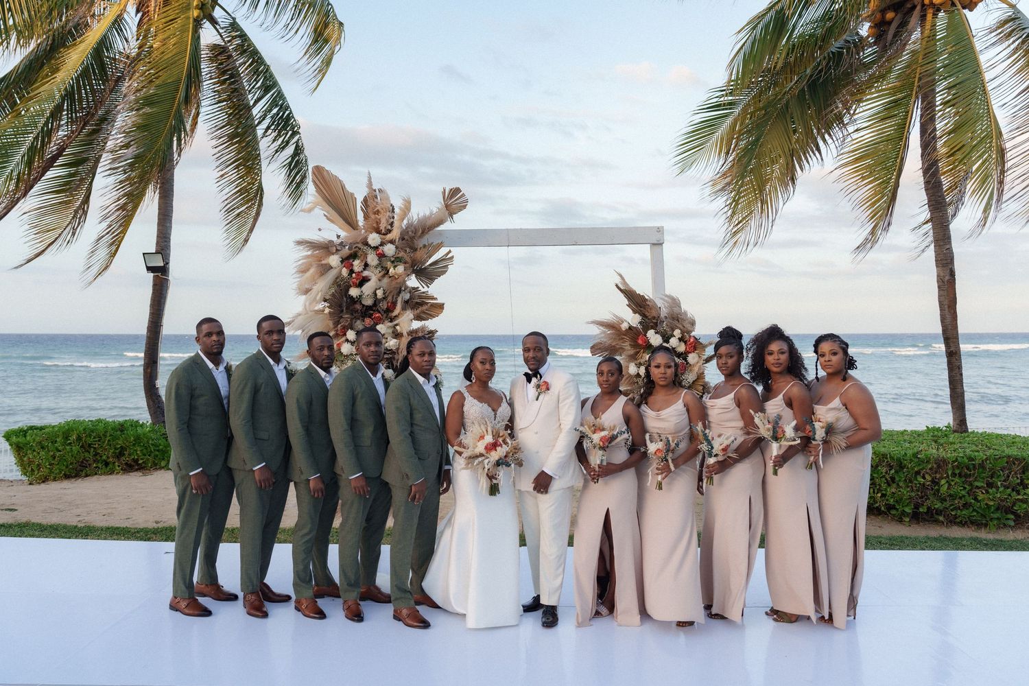 Wedding party in sage green and blush poses on oceanfront deck with palm trees.