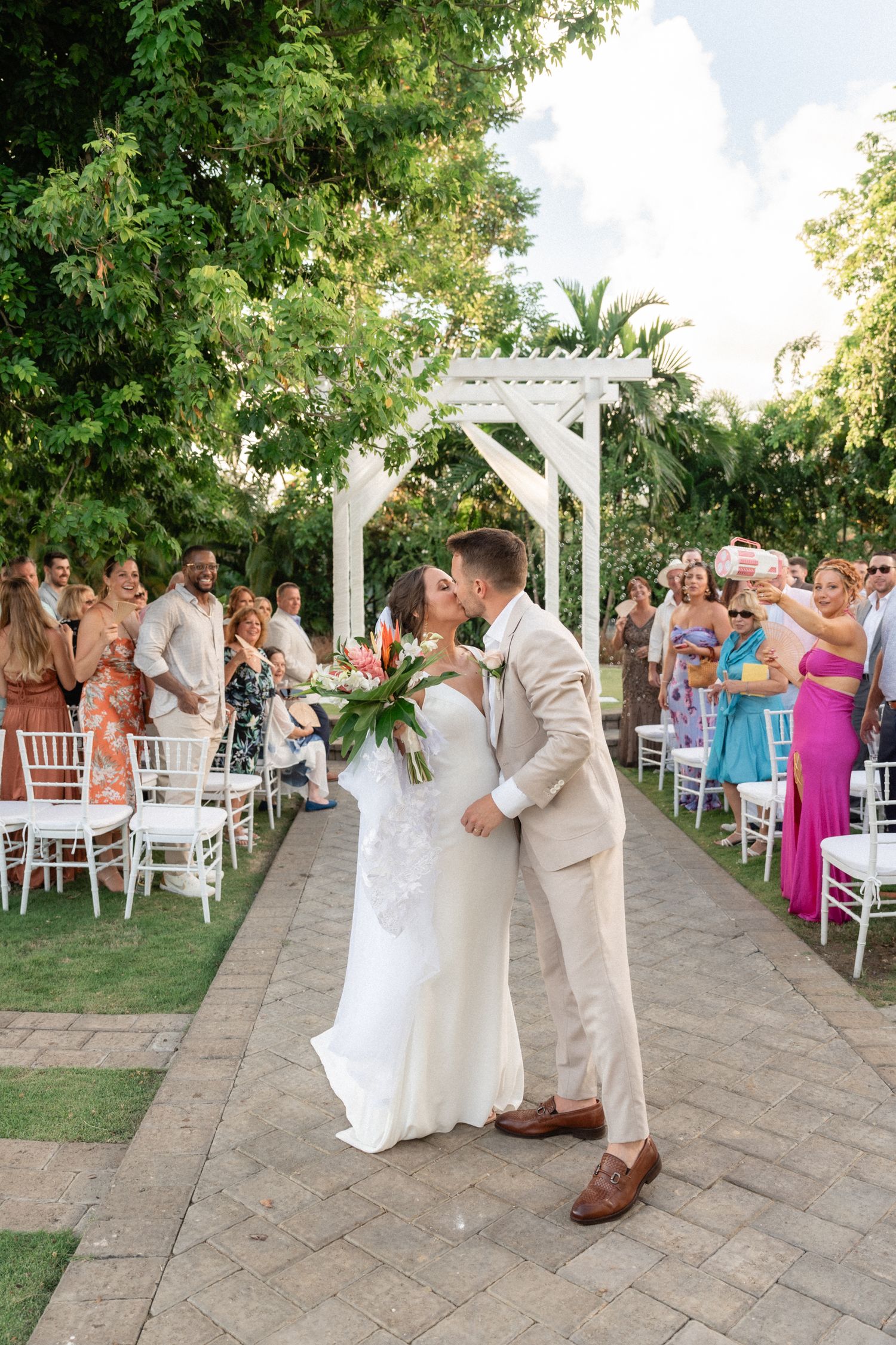 A wedding ceremony moment under a white pergola with guests seated on both sides.