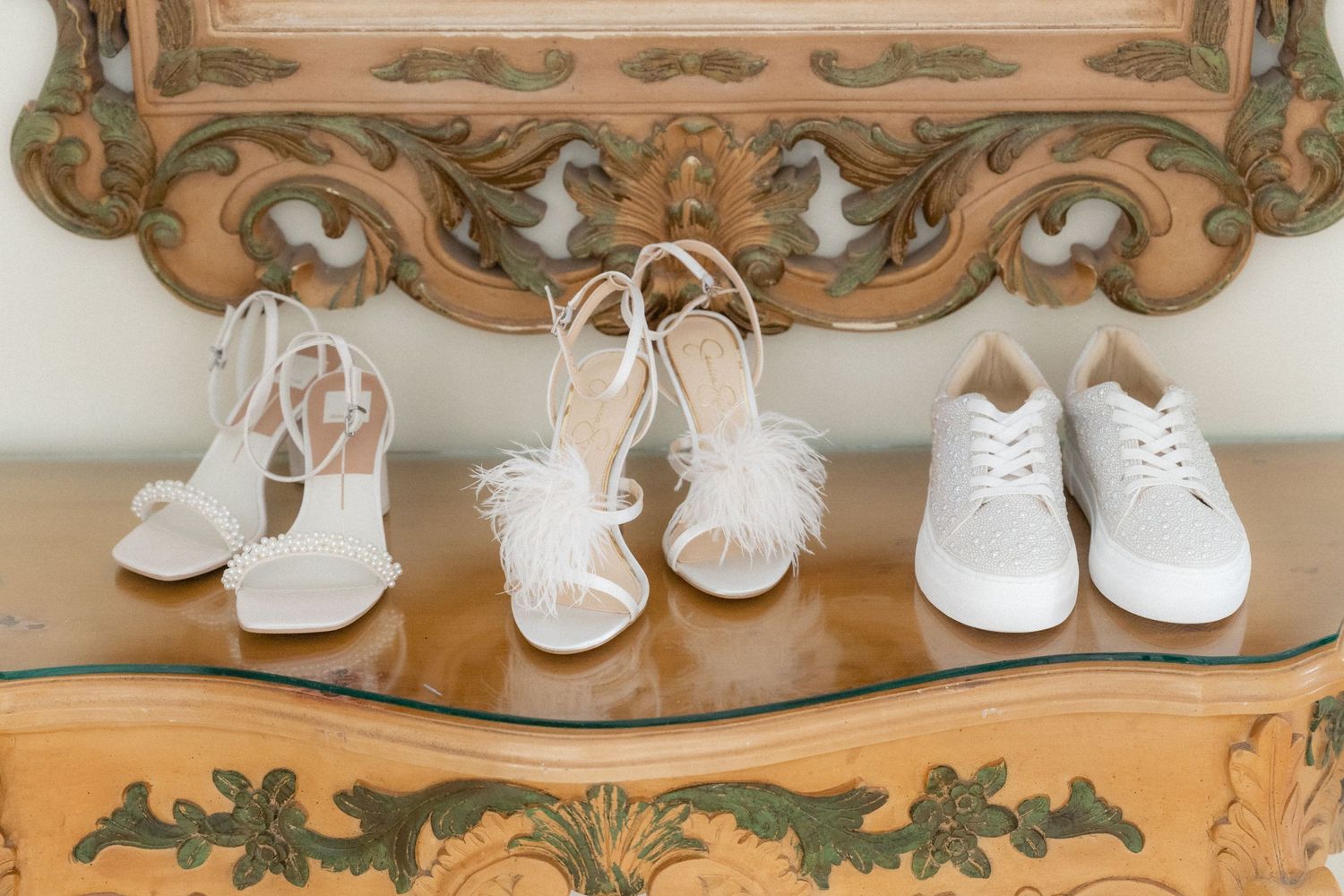 Three pairs of white wedding shoes displayed on an ornate vintage wooden console table.