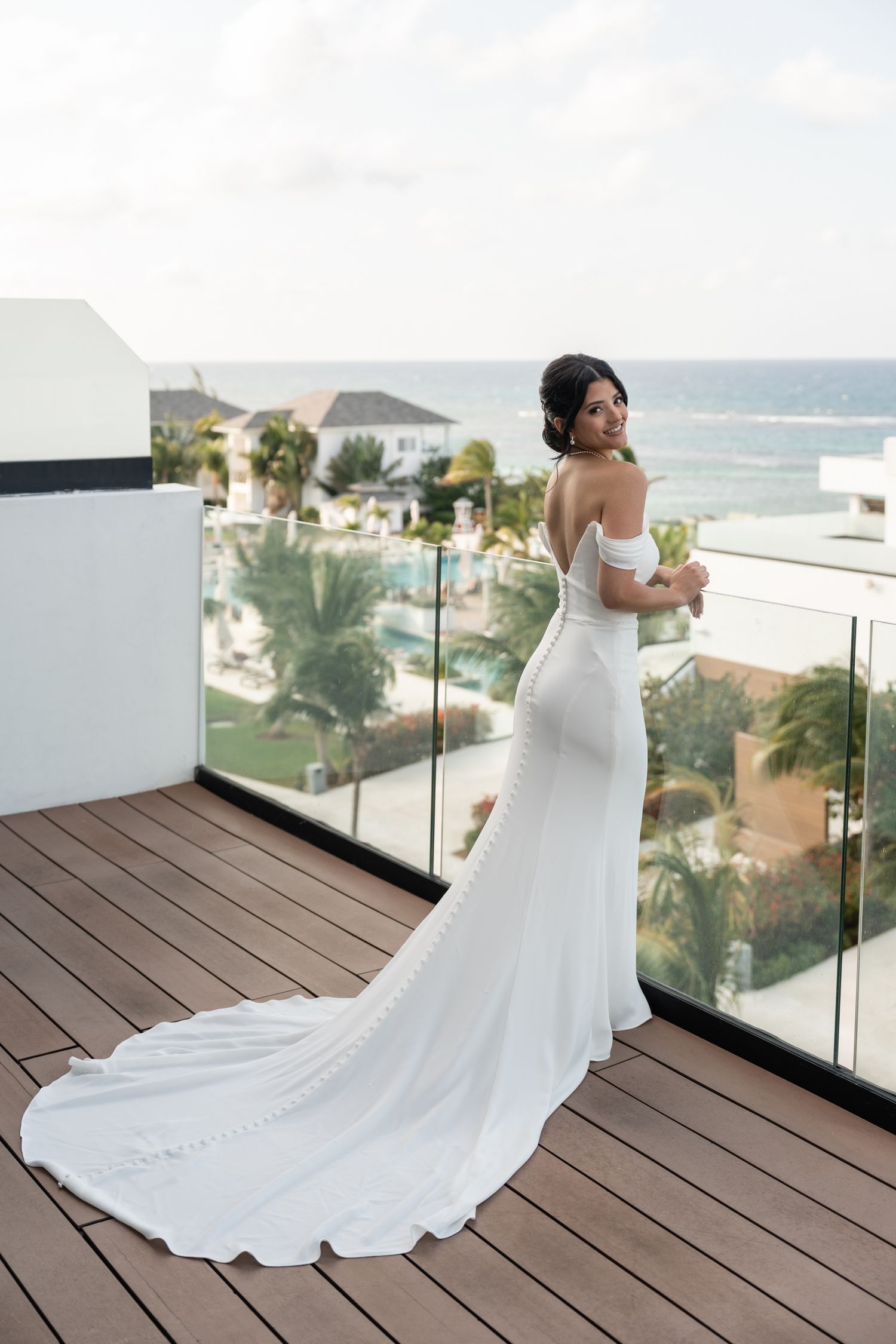 Bride in fitted white gown with train poses on oceanfront balcony deck.