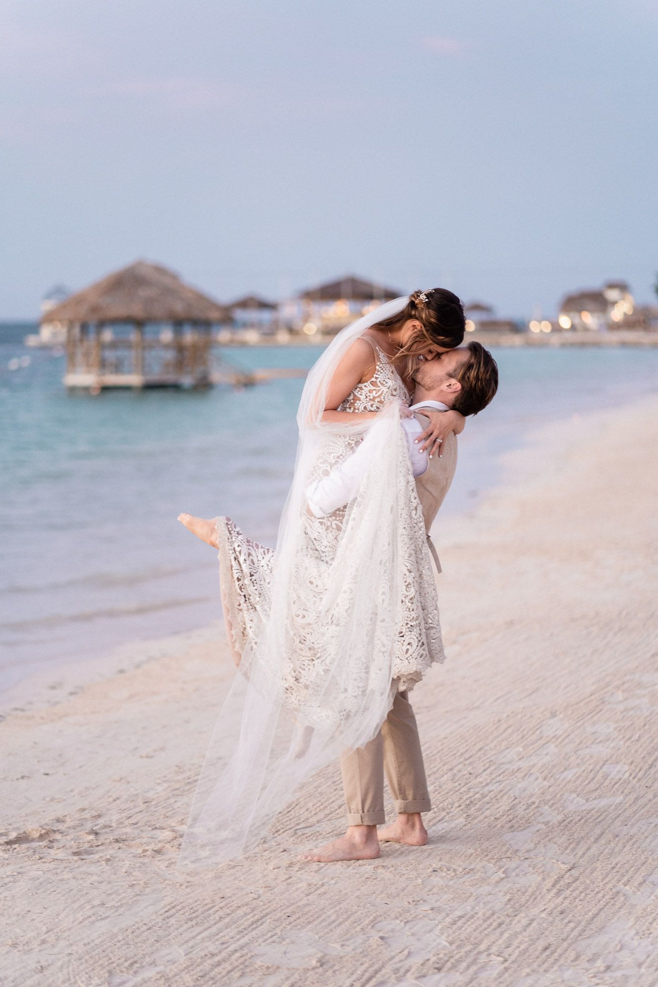 A romantic couple embraces during their beach wedding photoshoot with overwater bungalows visible.