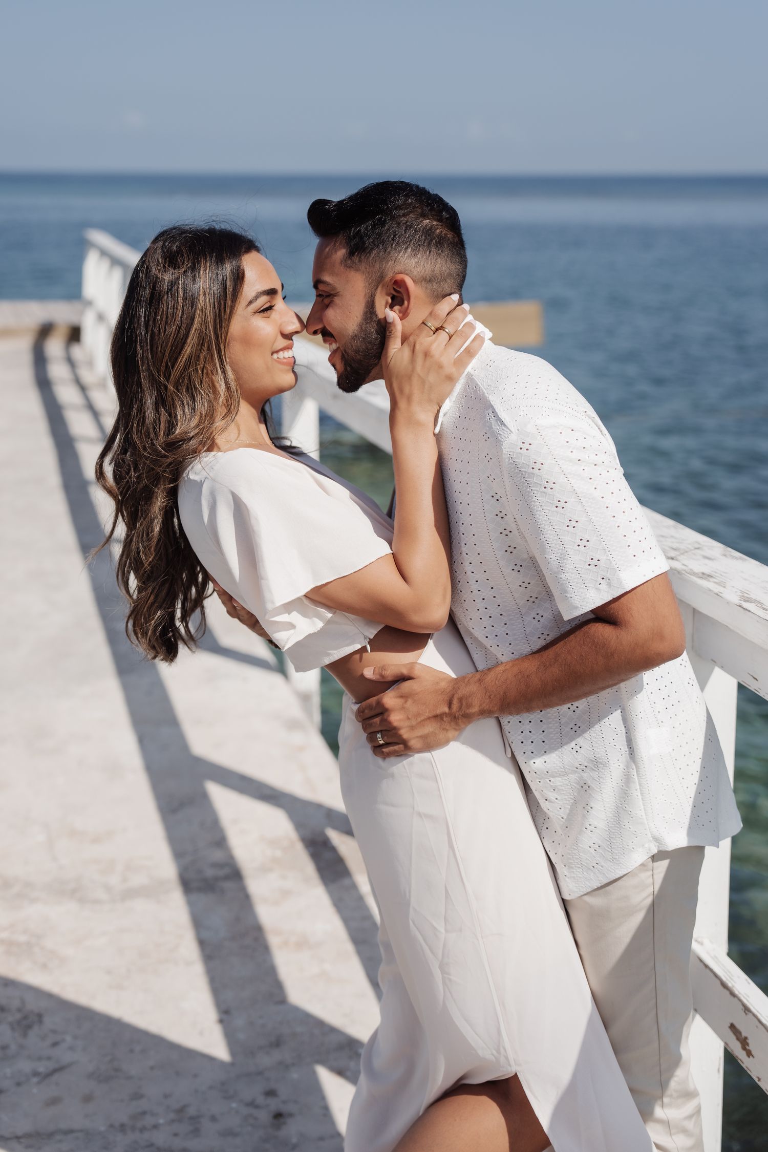 A romantic moment between a couple in white outfits on a pier overlooking the ocean.
