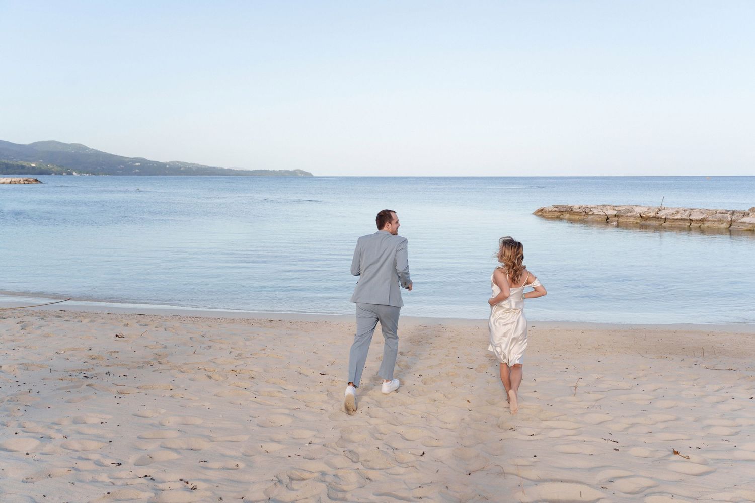 Couple running playfully together on a sandy beach with a stone pier in the background.