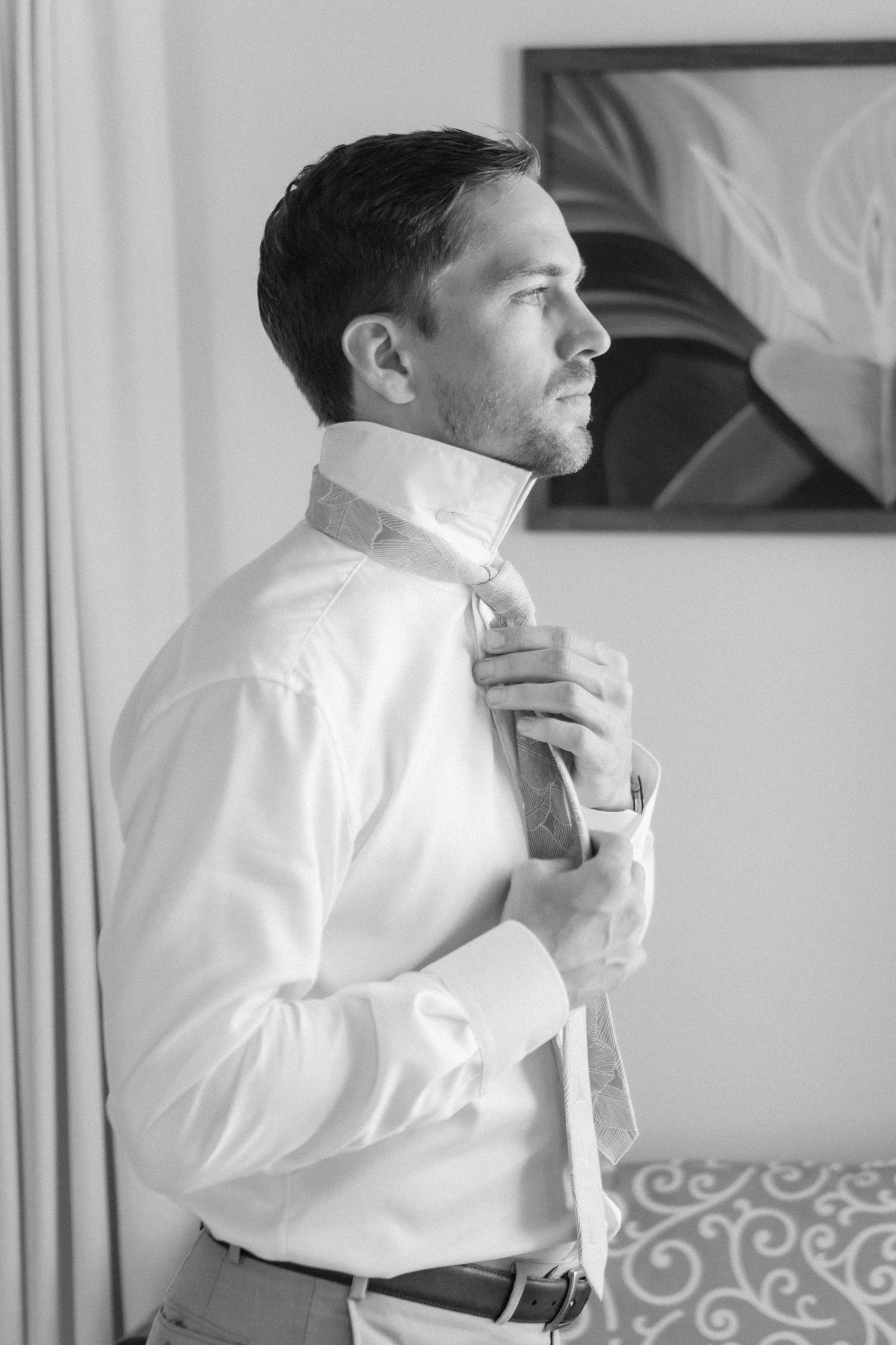 Groom in white shirt adjusting tie while getting ready in monochrome portrait.