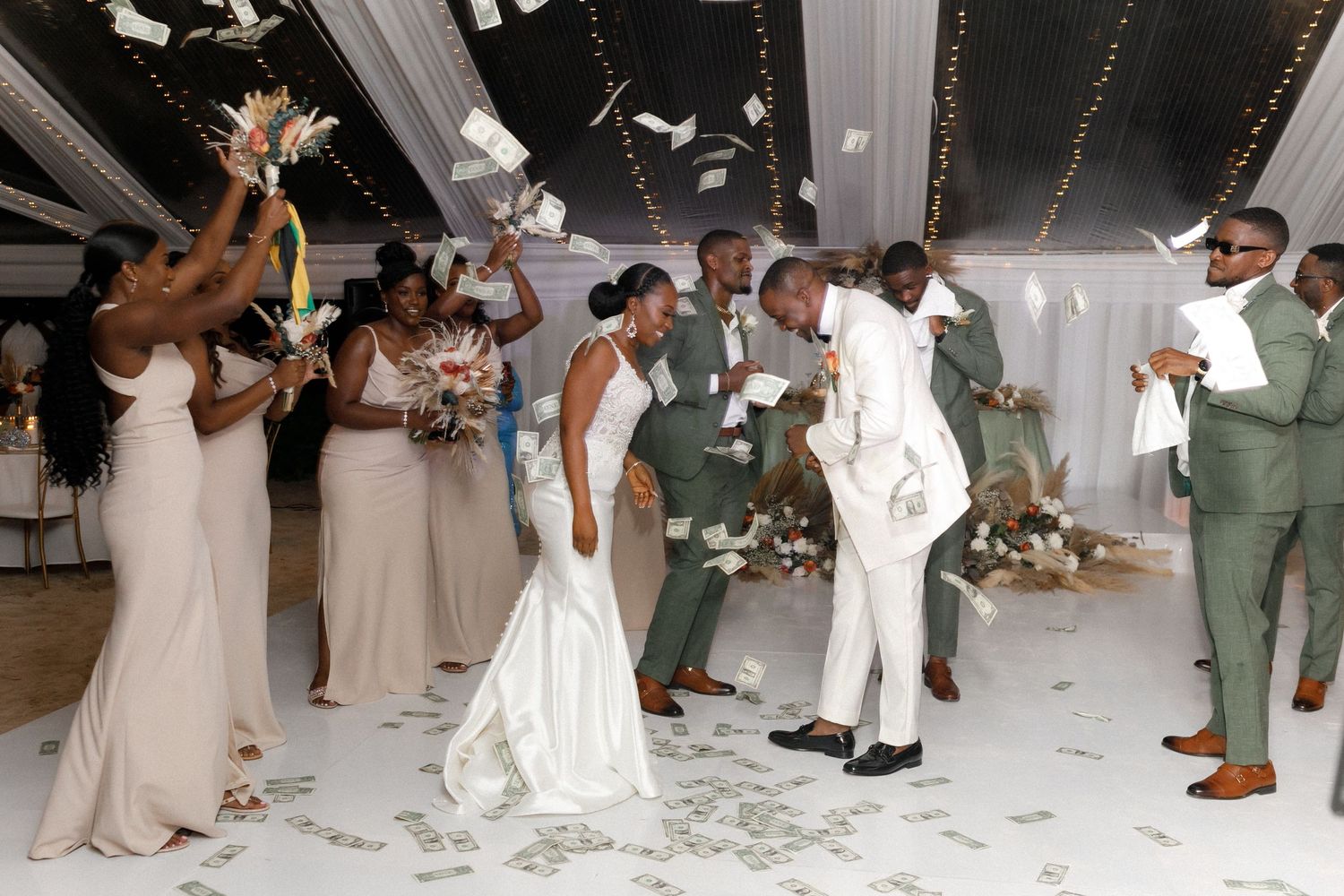 Wedding party dancing and celebrating on white dance floor under draped ceiling with string lights.