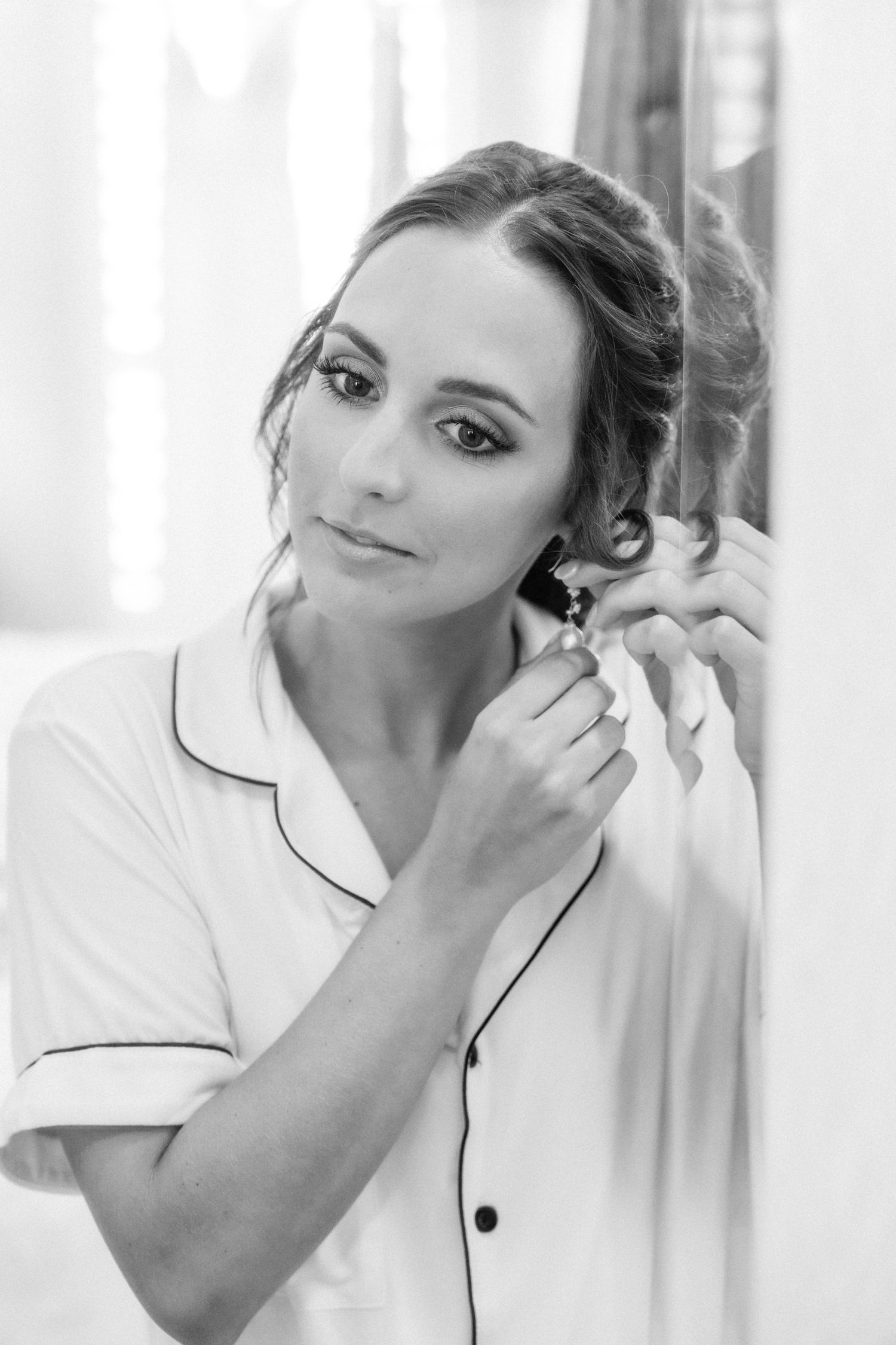 A black and white portrait shows a woman in a collared top putting in an earring.