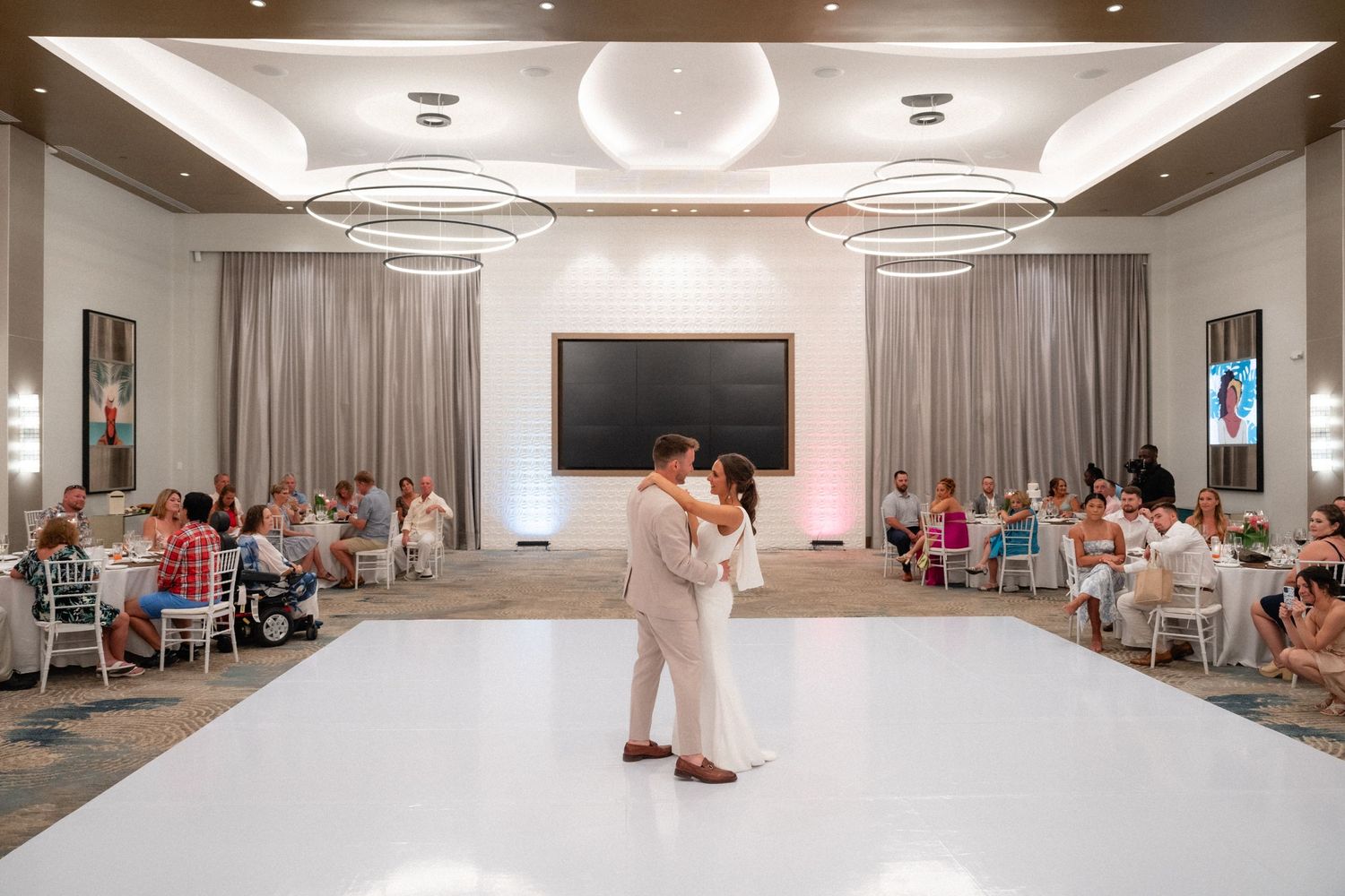 Wedding celebration in an elegant ballroom with white decor and modern lighting.