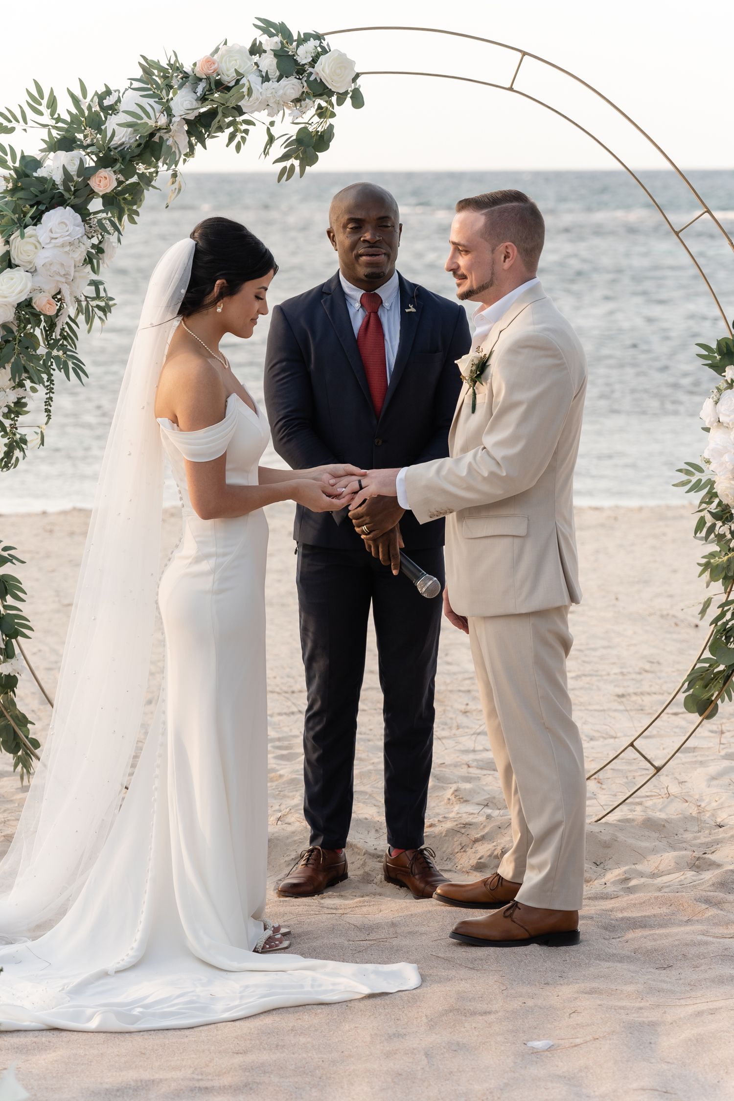 A wedding ceremony takes place on the beach under an elegant circular arch adorned with white flowers and greenery.