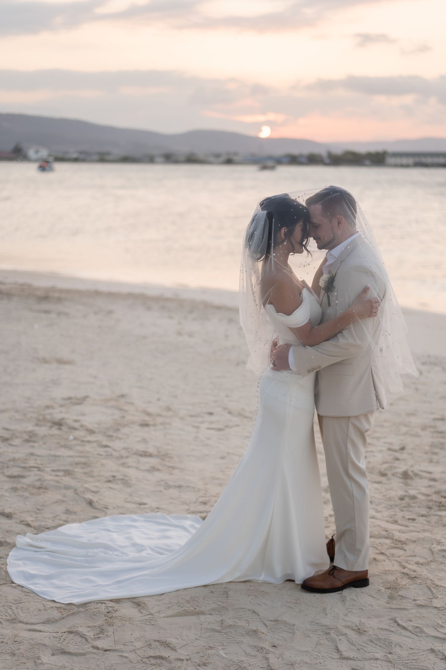 Newlyweds share intimate moment on beach at sunset with flowing wedding dress train.