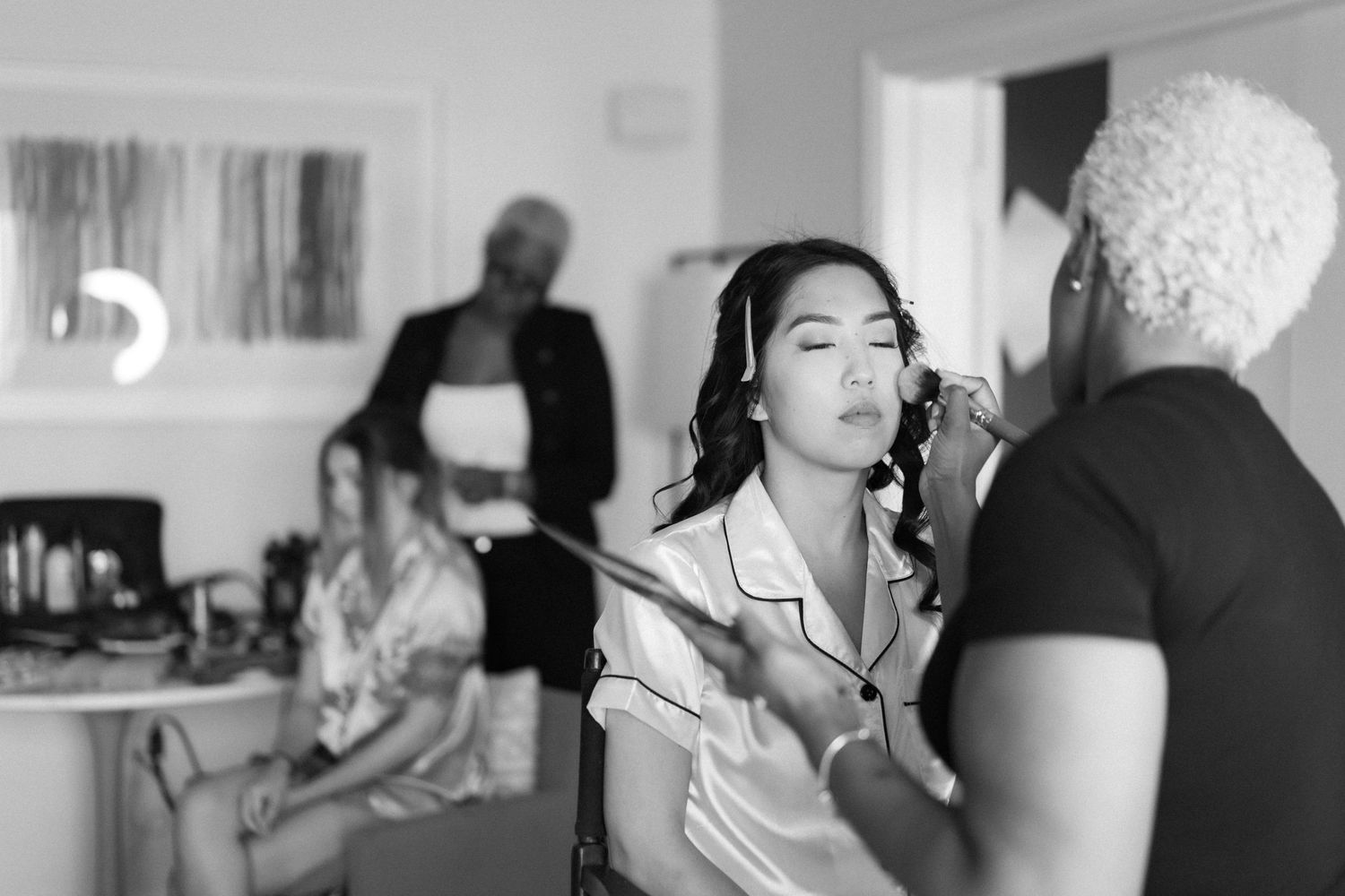 Black and white photo of bride getting professional makeup applied during wedding preparation.