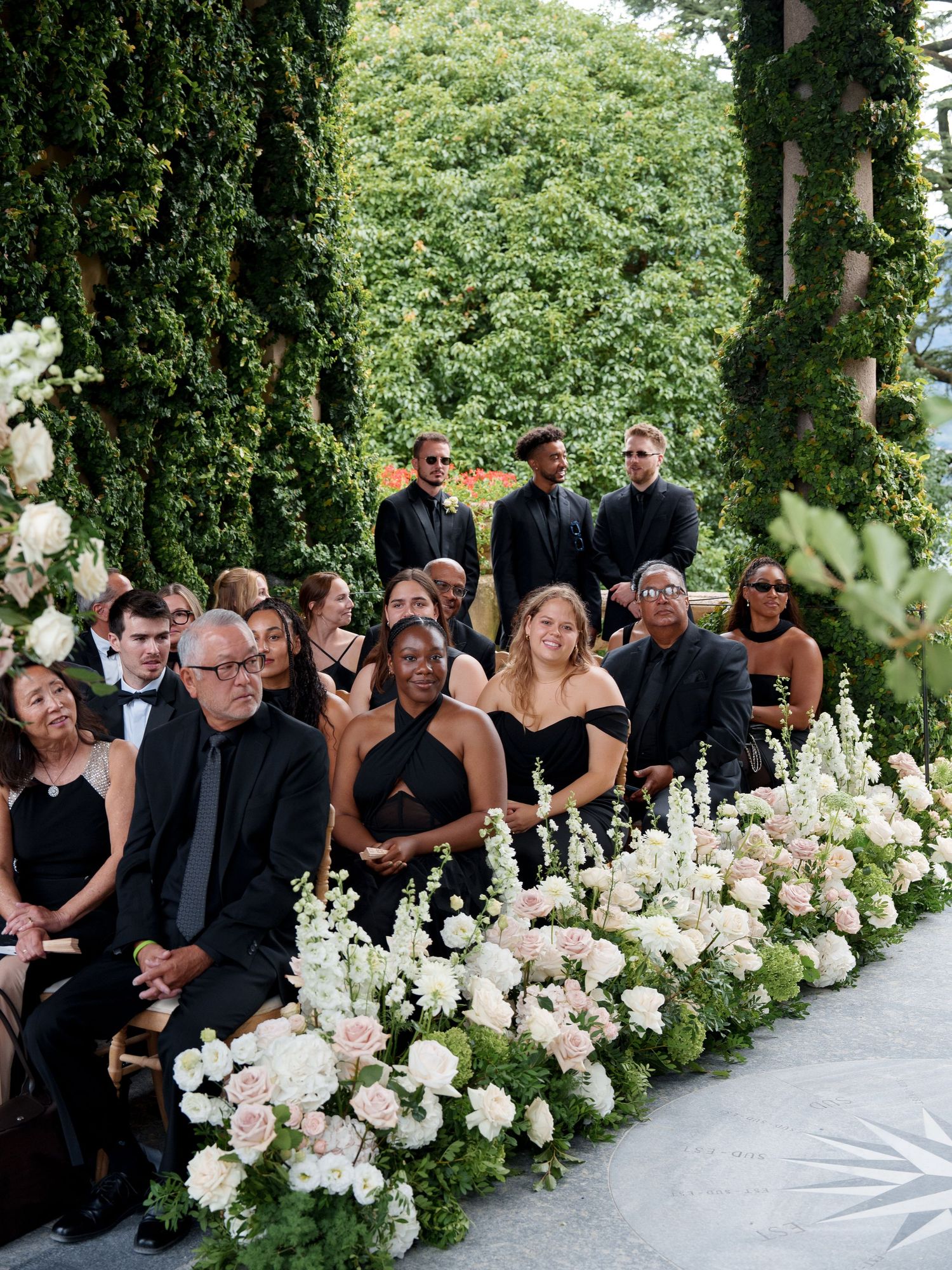 Wedding guests dressed in black formal attire seated in rows with elegant white floral arrangements lining the aisle.