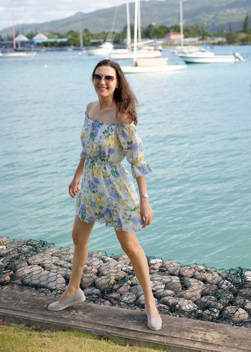 Woman in floral blue romper poses playfully on rocky shoreline with boats and ocean in background.