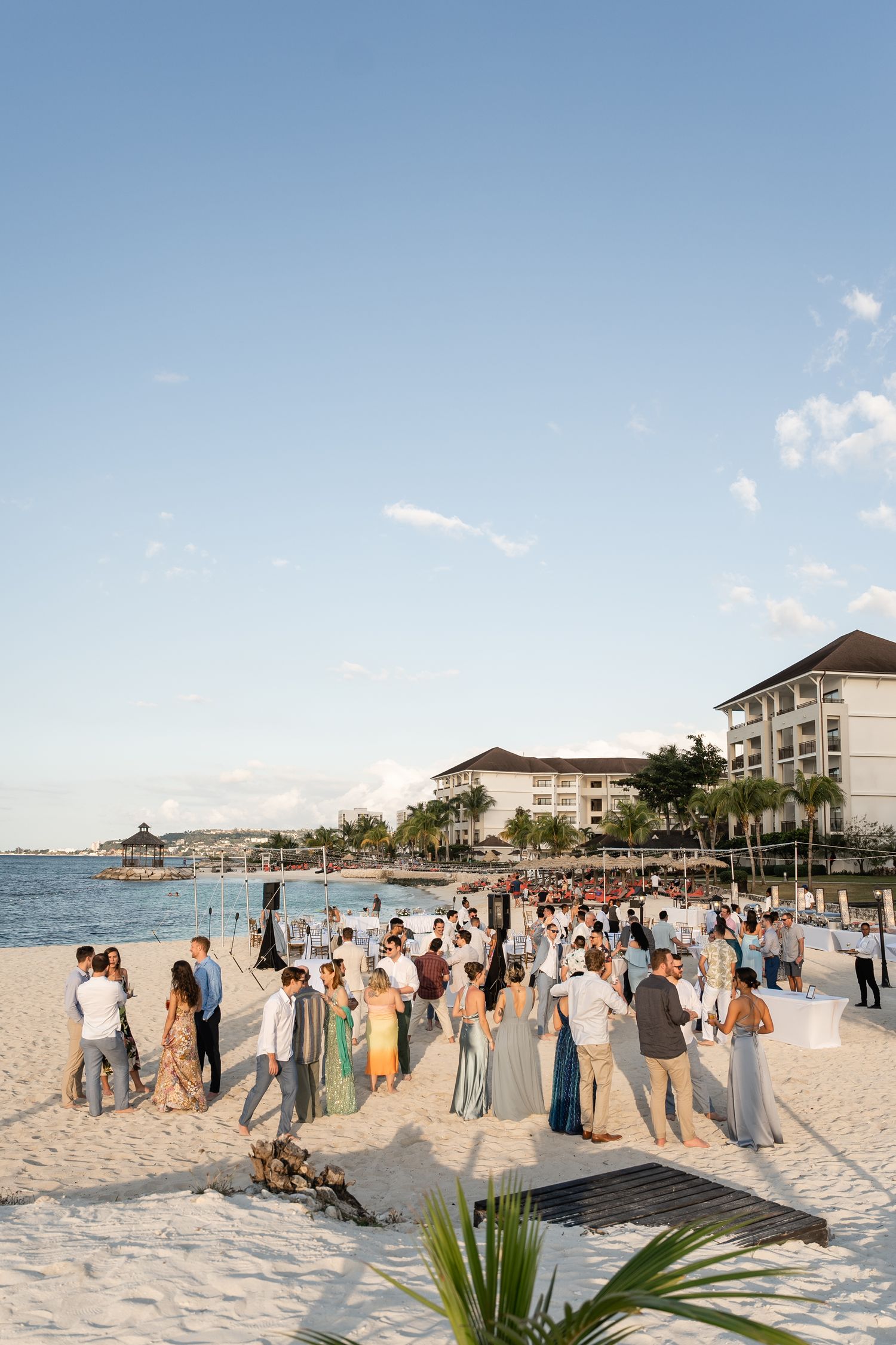 Wedding guests gathered on resort beach at sunset with hotel buildings in background.