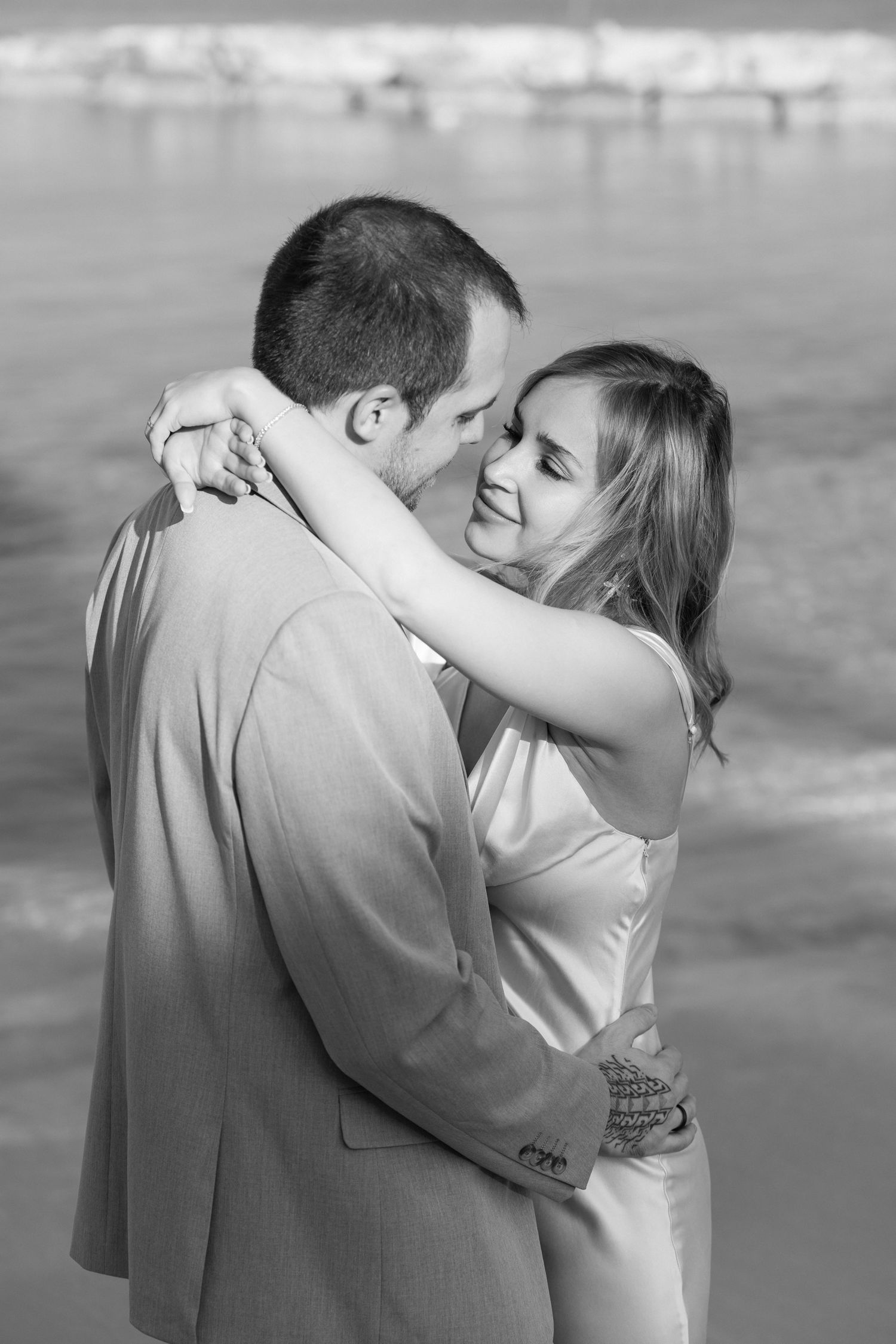 Black and white portrait of couple embracing on beach with water in background.