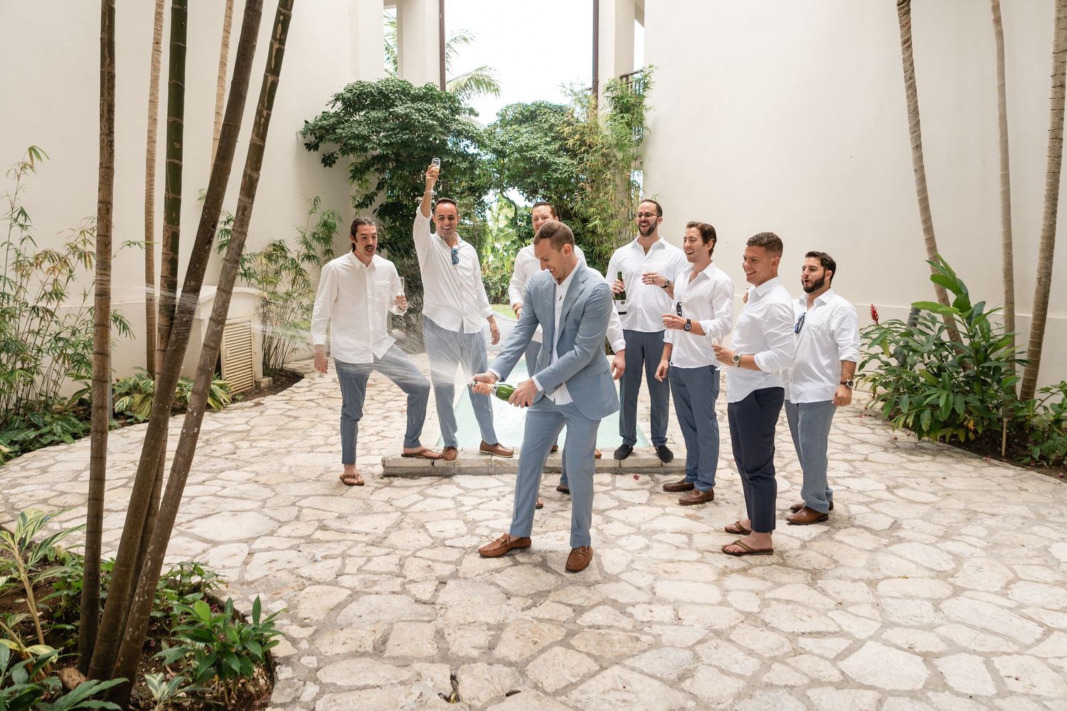 Wedding party gathered in tropical courtyard wearing coordinated white and gray outfits.