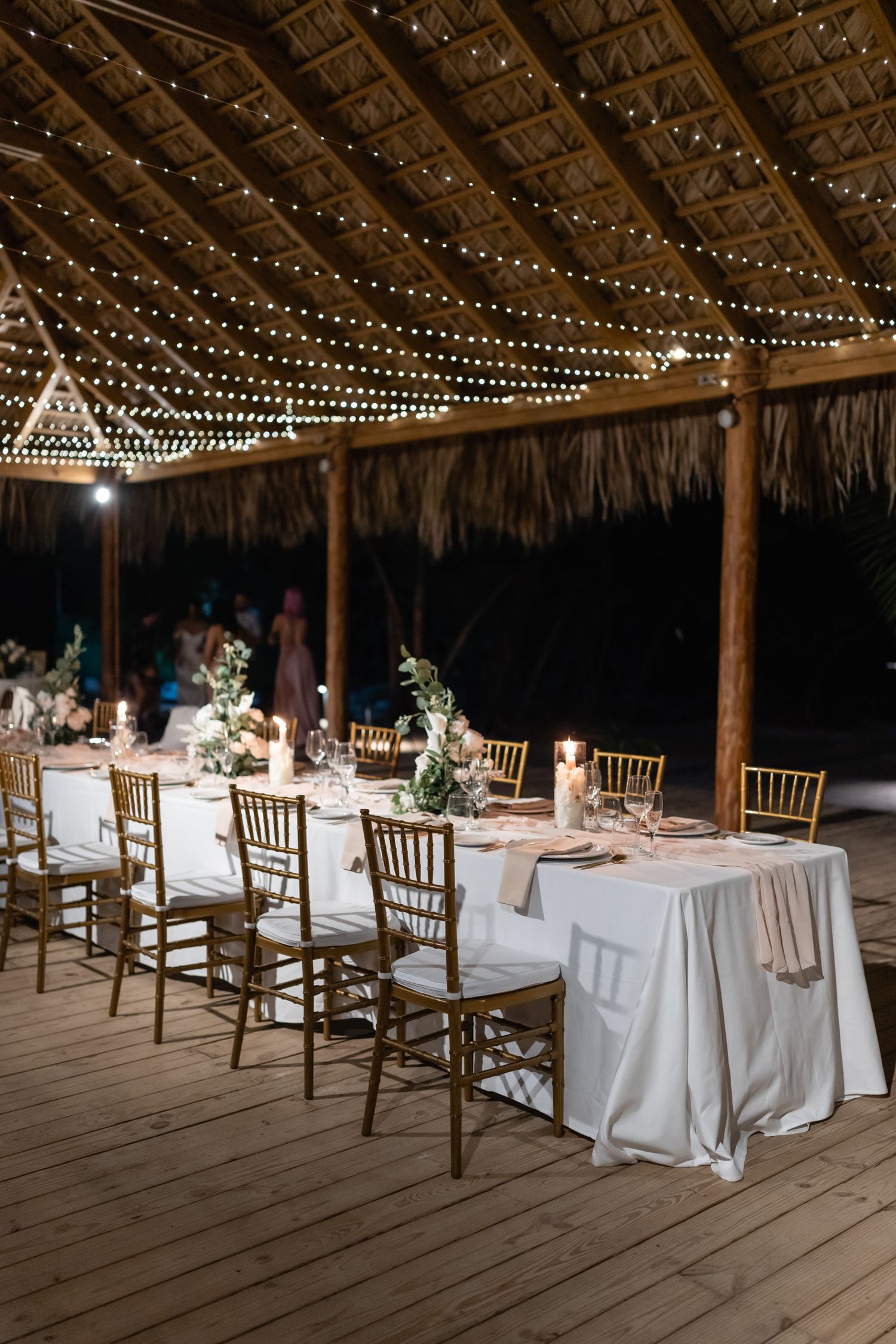 Romantic reception setup under thatched roof with string lights and elegant table settings.
