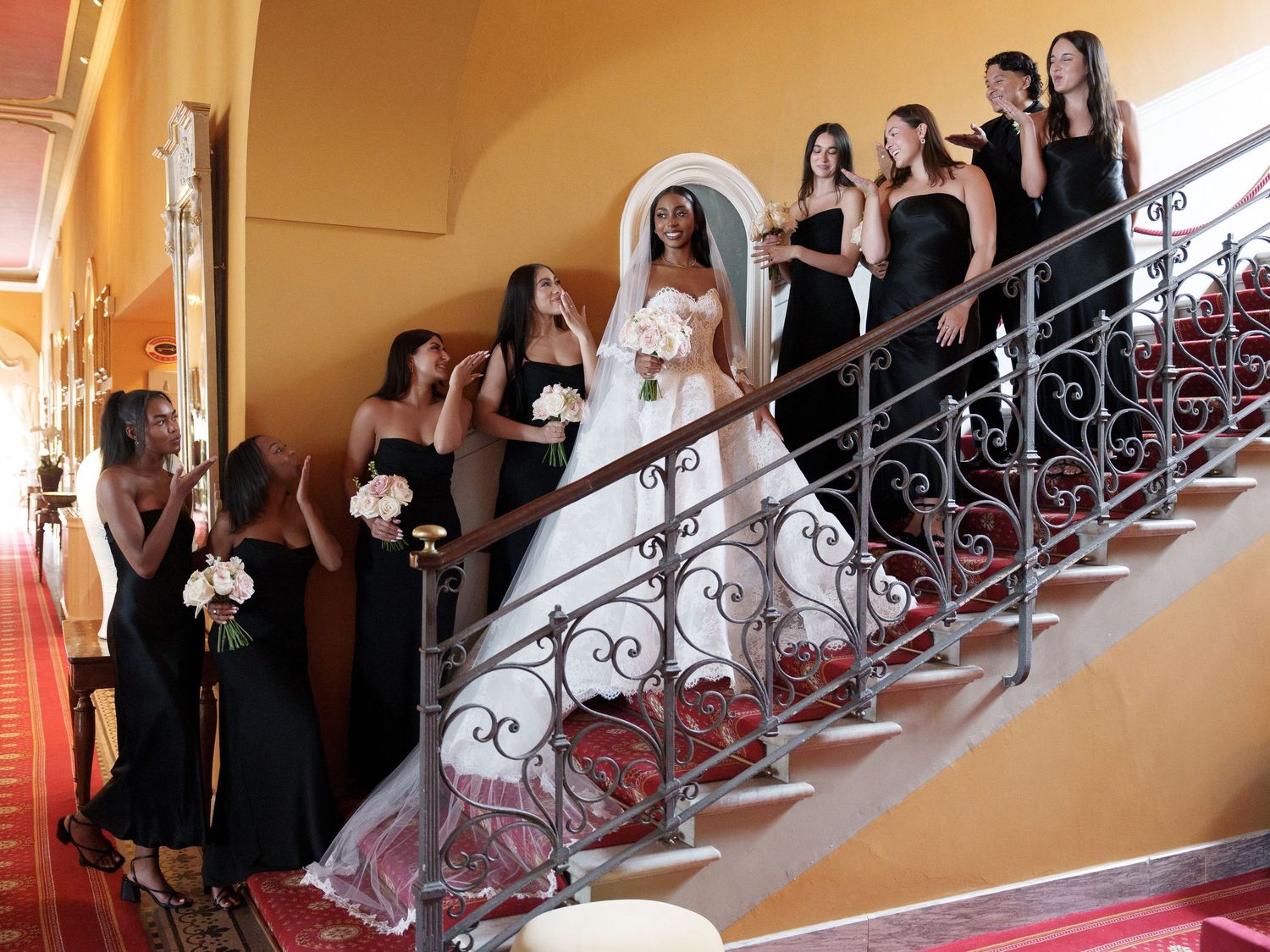 Wedding party members in black dresses pose along an elegant staircase with ornate iron railings.