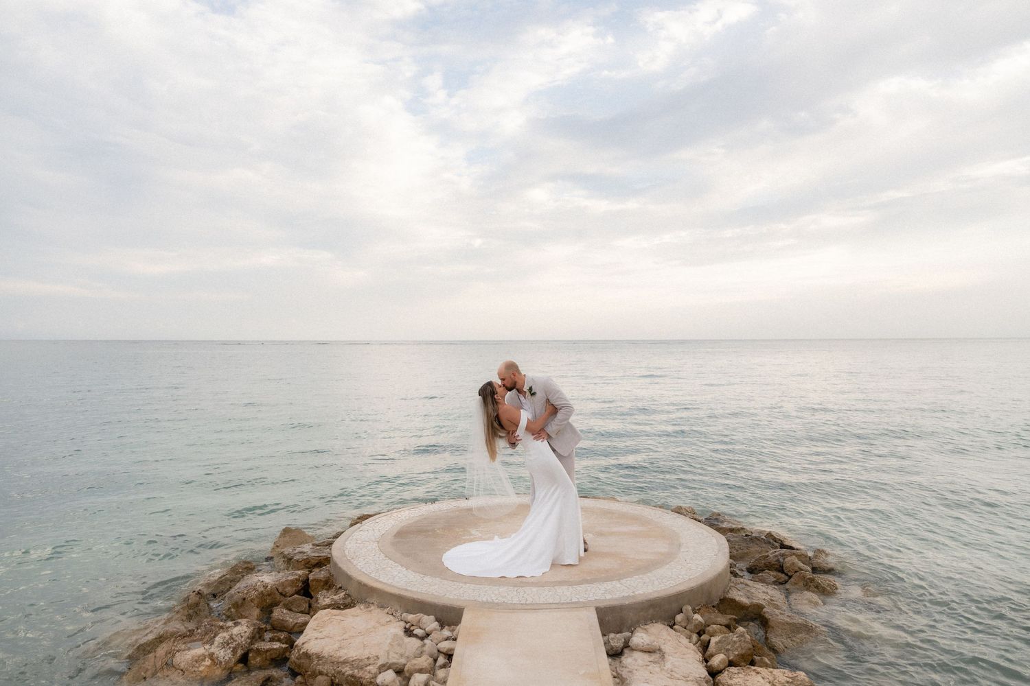 Romantic wedding photo on circular stone platform overlooking ocean at sunset.
