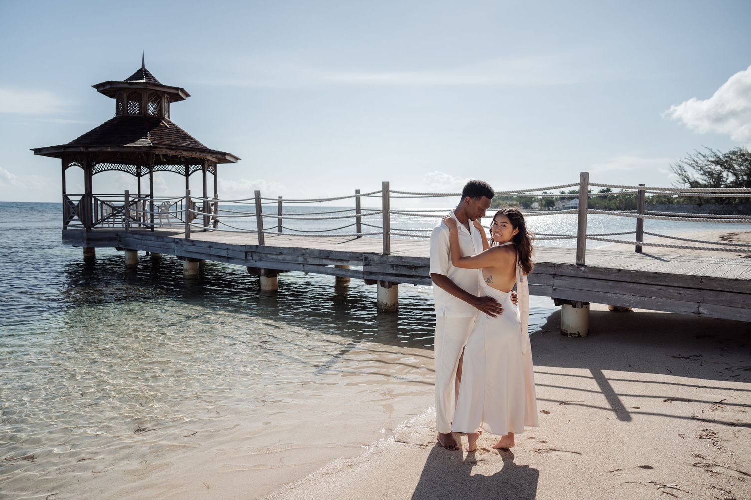 A couple embraces on a wooden pier with a gazebo and tropical ocean backdrop.