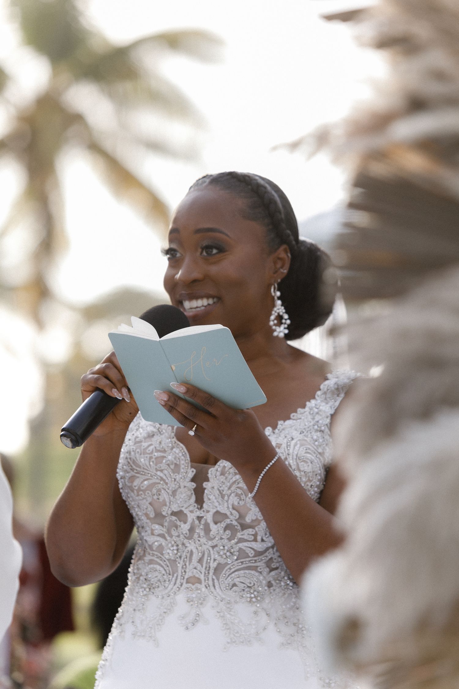 Bride in lace wedding dress reading vows from a blue book during outdoor ceremony.