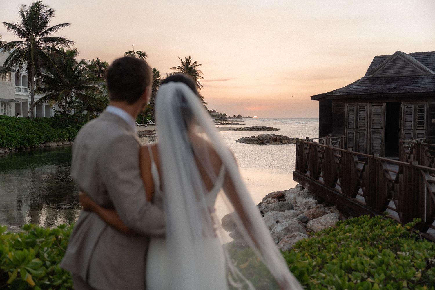 A couple in wedding attire watches the sunset over a tropical waterfront with palm trees and rustic buildings.