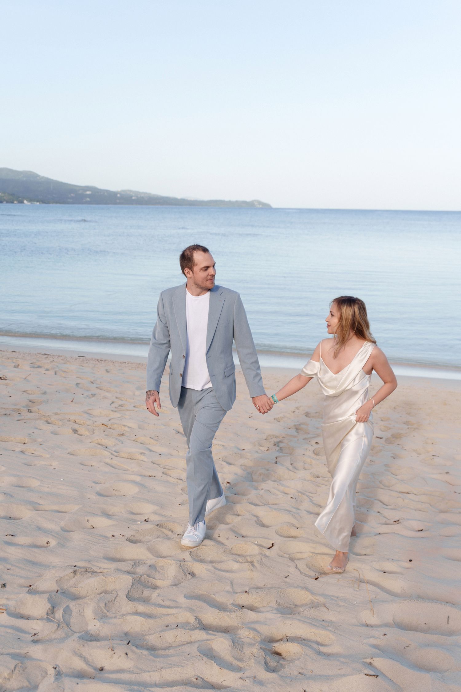 Couple walking hand in hand on a beach at sunset, wearing formal attire and white sneakers.