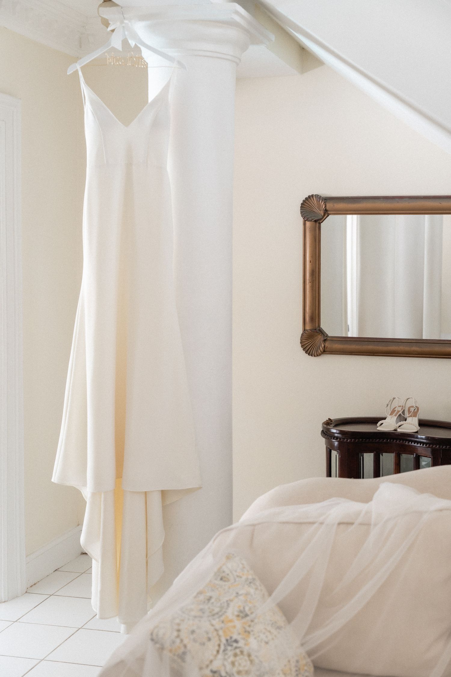 Elegant white wedding dress hanging in a bright room with a mirror and veil.