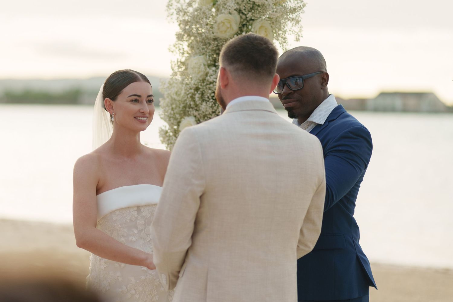 A beachside wedding ceremony takes place with a couple and officiant standing together at sunset.