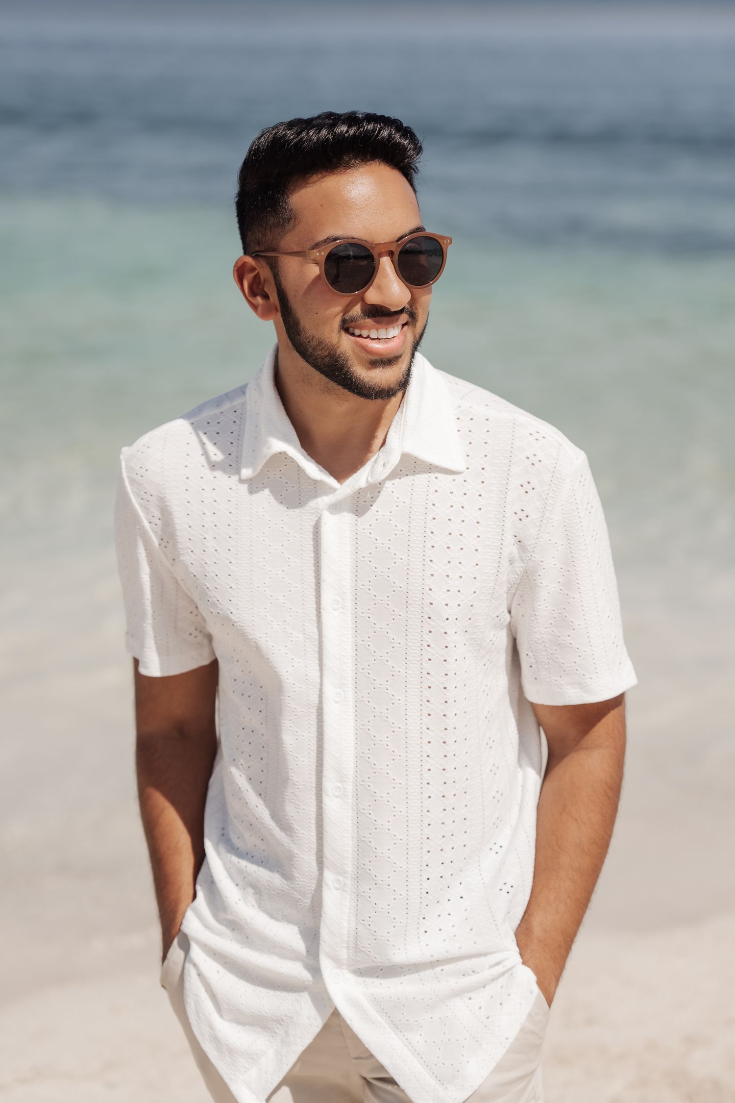 Man in white shirt and sunglasses smiles on tropical beach.