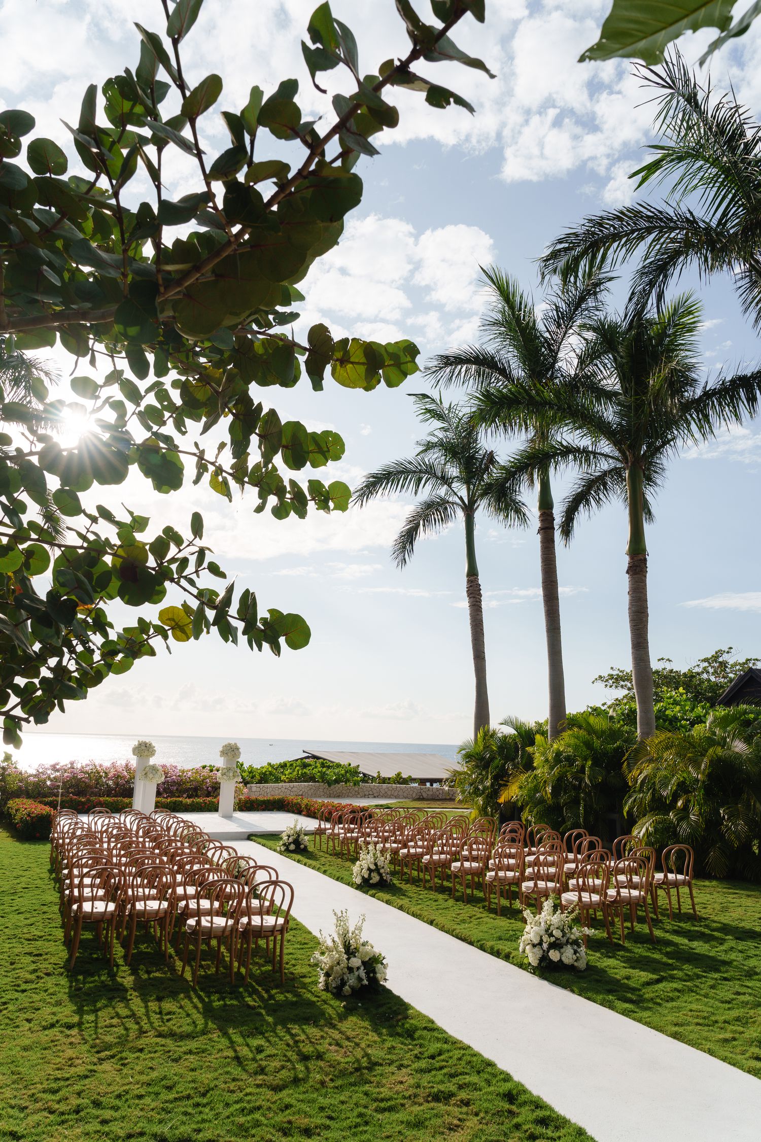 Outdoor wedding ceremony setup with rows of wooden chairs along white aisle runner framed by tall palm trees and ocean view.