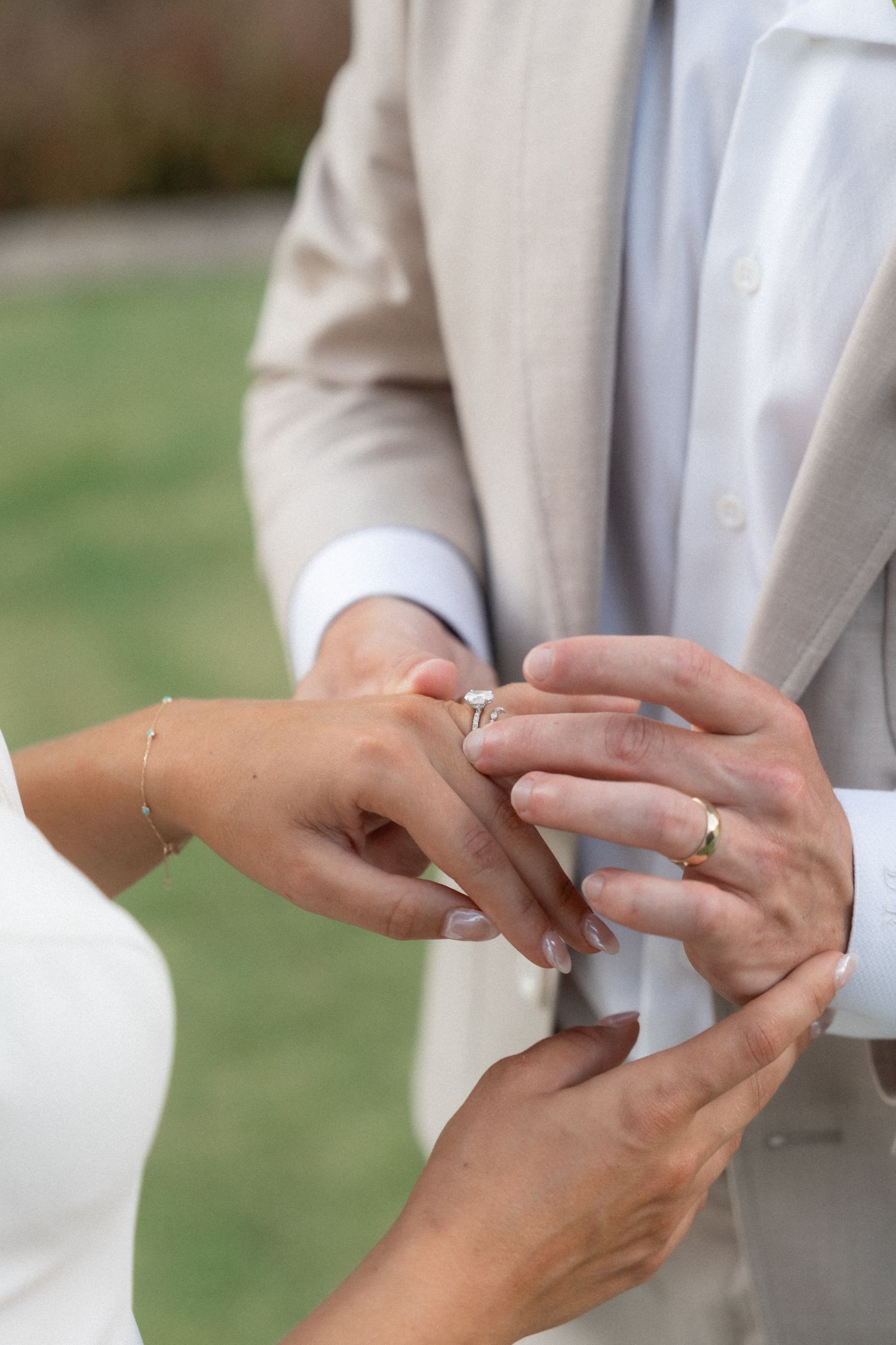 Close-up of hands exchanging wedding rings during ceremony, with groom wearing light beige suit.