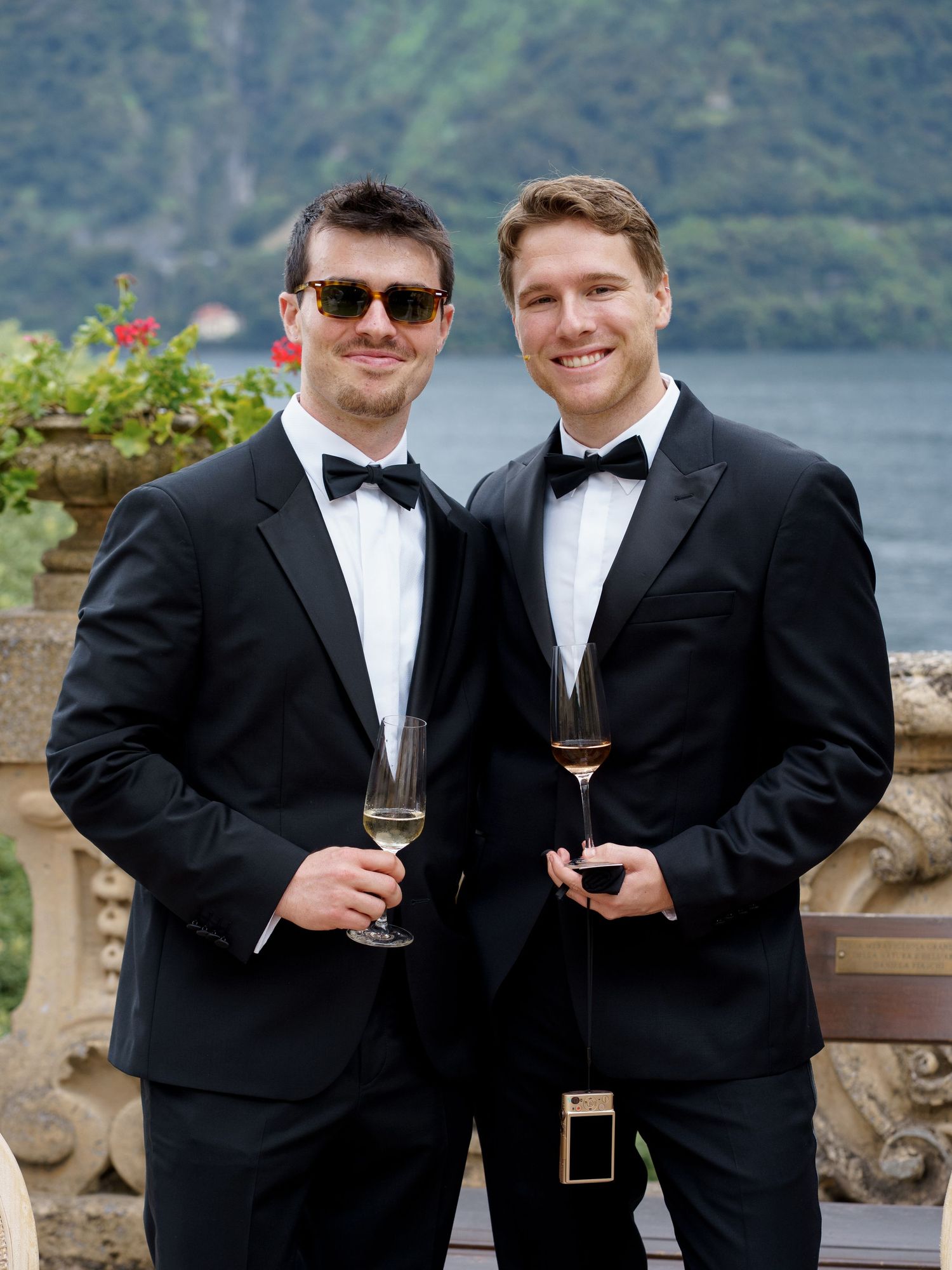 Two people in black tuxedos and bow ties hold wine glasses while posing outdoors against a scenic waterfront view.