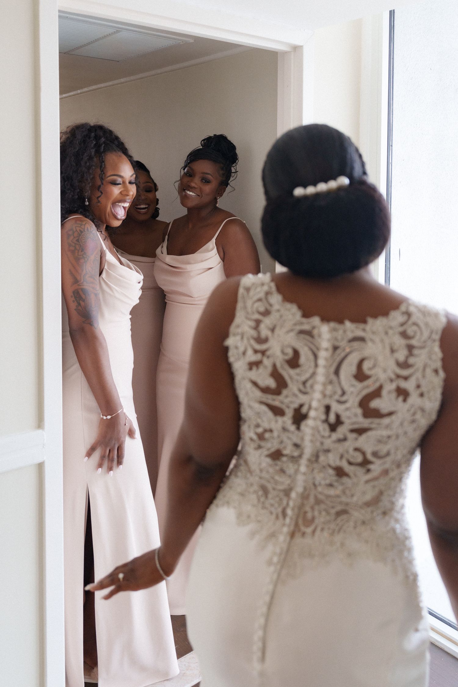 Bridesmaids in pink dresses reacting with joy to seeing the bride in her lace wedding gown.