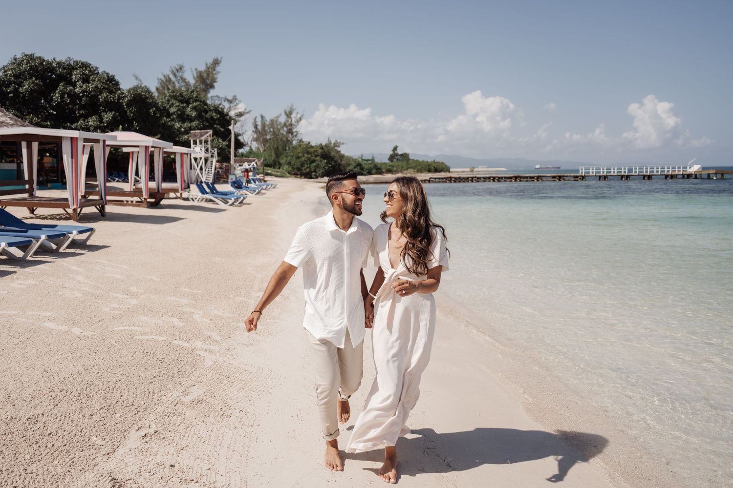 Couple walking hand in hand along a tropical beach with lounge chairs in background.