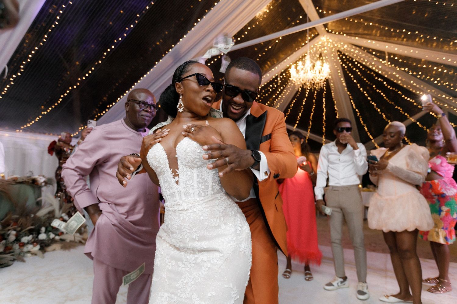 Wedding celebration in full swing with guests dancing under crystal chandelier and string lights.